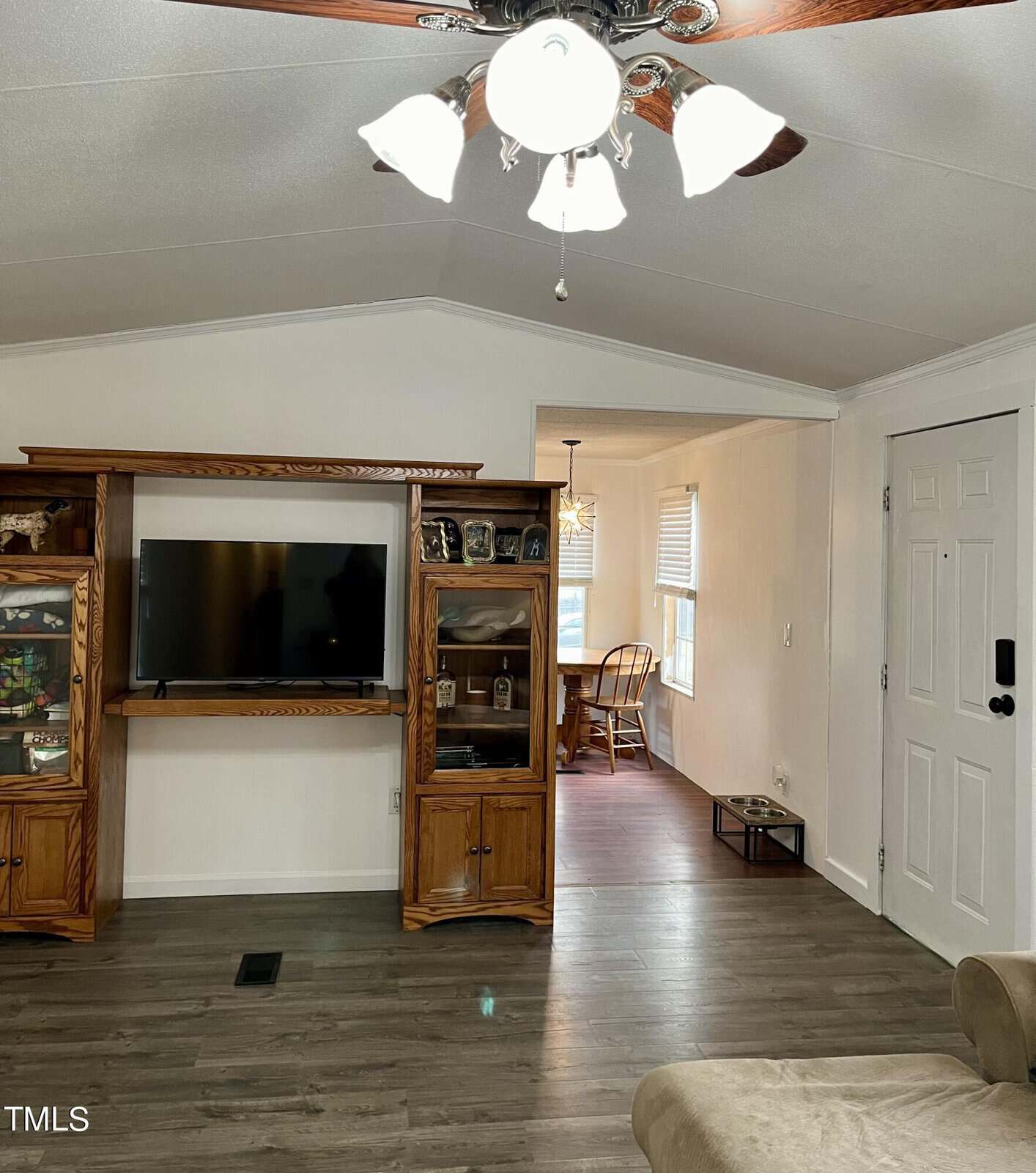 A cozy living room with wood laminate flooring, a ceiling fan with lights, and a TV mounted on a wooden entertainment center with glass cabinets on either side. The entry door is visible to the right, and a dining area with a wooden chair is seen through an open doorway.