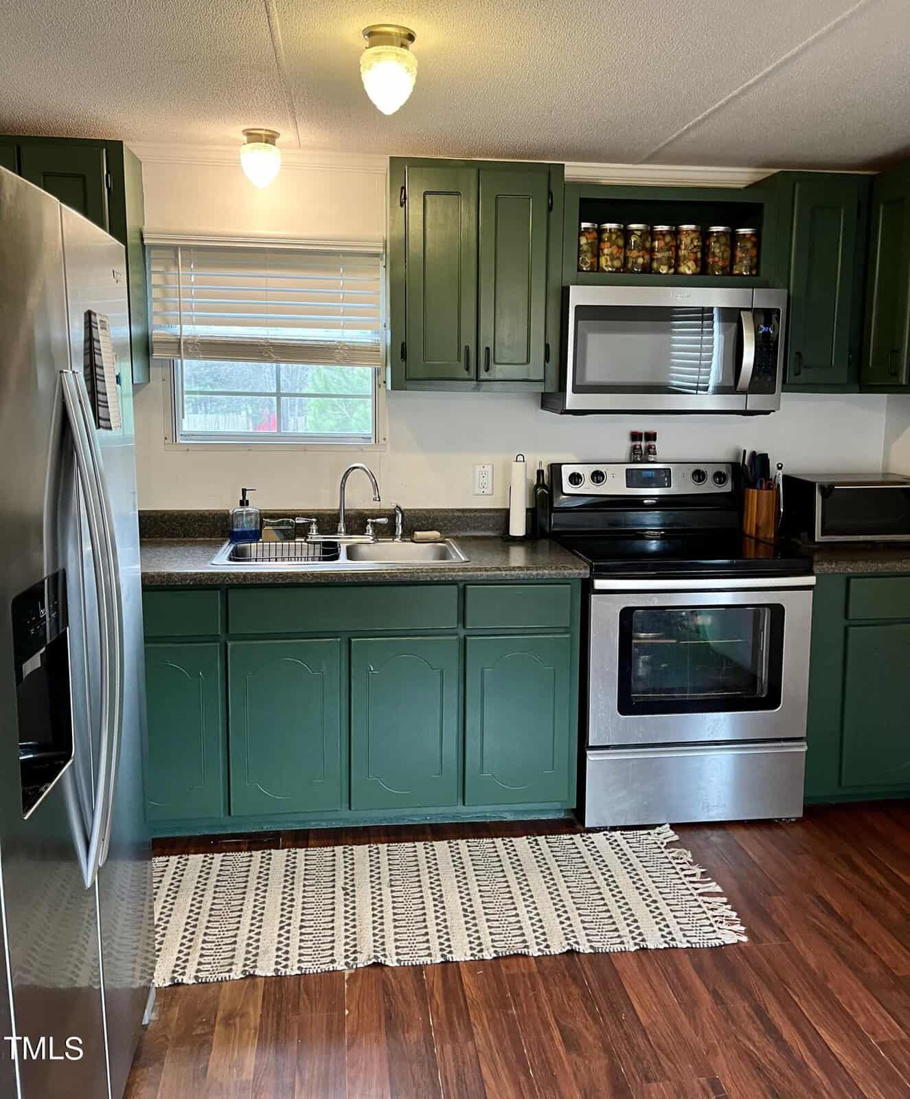 A kitchen with green cabinets, stainless steel appliances including a refrigerator, oven, microwave, and dishwasher. The countertop holds jars above the microwave and a dish rack near the sink. A patterned rug lies on the wooden floor in front of the sink.