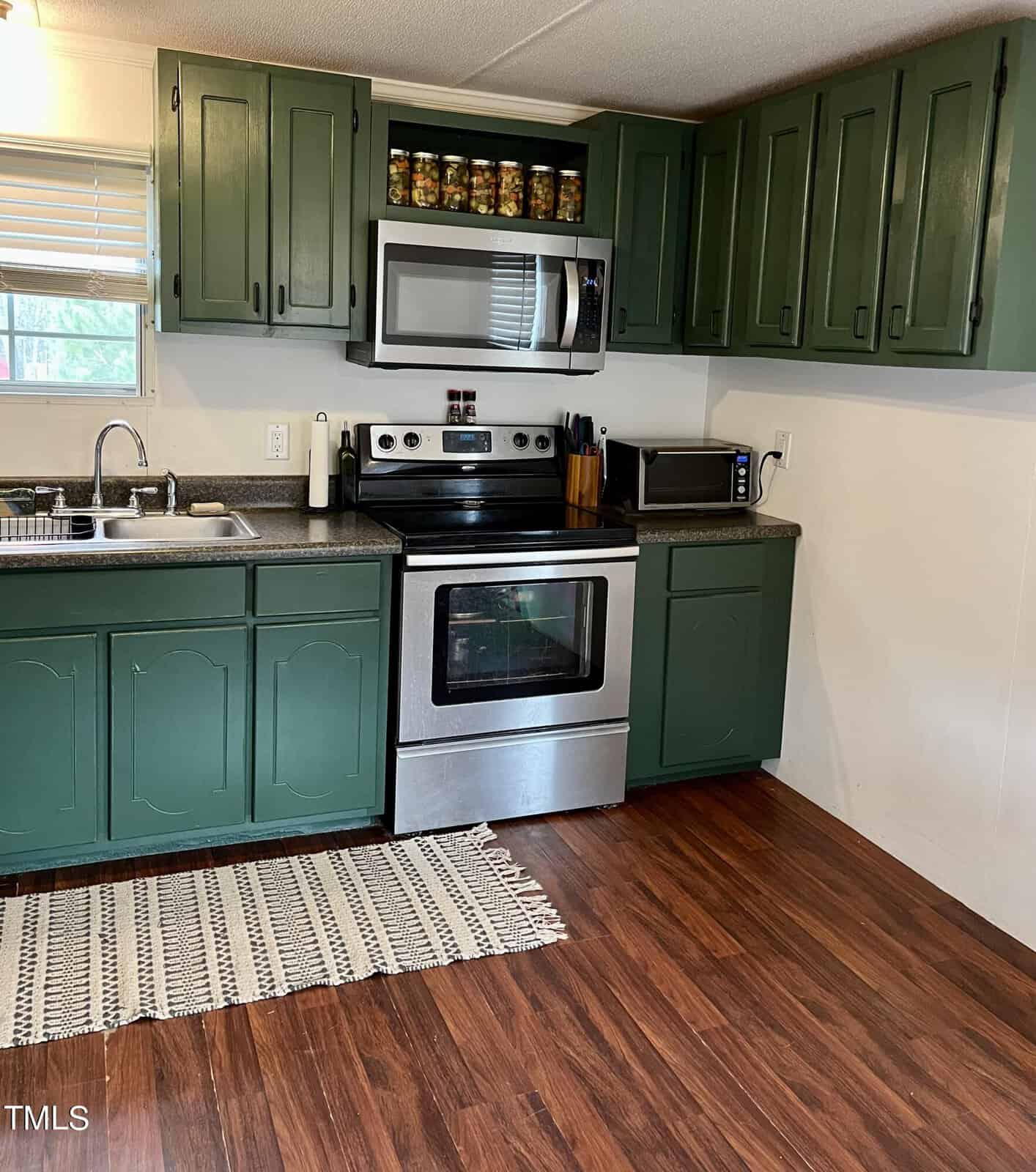 A kitchen with dark green cabinets, a stainless steel stove, microwave, and a small black toaster oven on the counter. Above the stove are glass jars stored on shelves. There is a sink on the left, and a striped rug on the wooden floor.