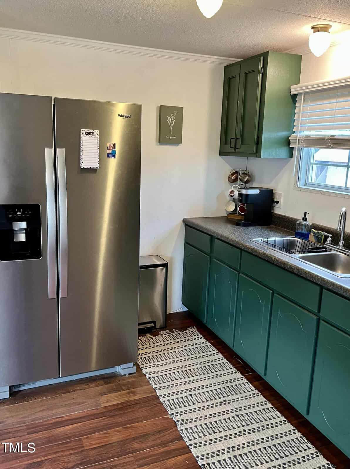 A kitchen with green lower cabinets and a large stainless-steel refrigerator. The countertop holds a coffee maker and a spice rack, and there's a dual sink below a window with blinds. A patterned rug lies on the wooden floor, and a small wall decoration hangs nearby.
