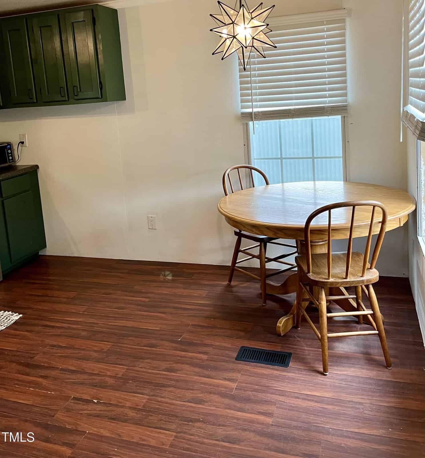 A small dining area featuring a round wooden table with two matching chairs on a hardwood floor. Above the table hangs a star-shaped light fixture. The room has green cabinets on the left and two large windows with white blinds.