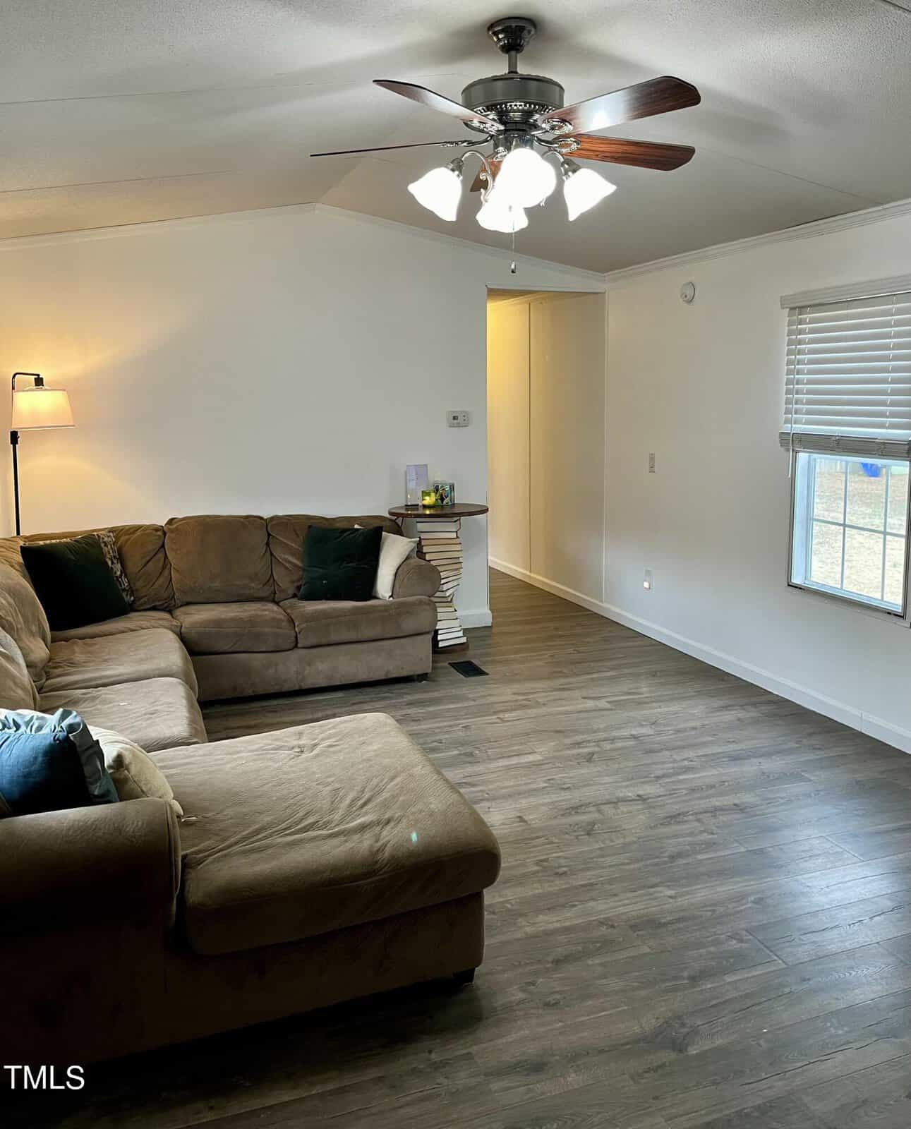 A spacious living room with wooden flooring, a large brown sectional sofa, a floor lamp, and a ceiling fan with lights. The room has white walls, a window with blinds, and a small side table near the entrance. Natural light illuminates the space.