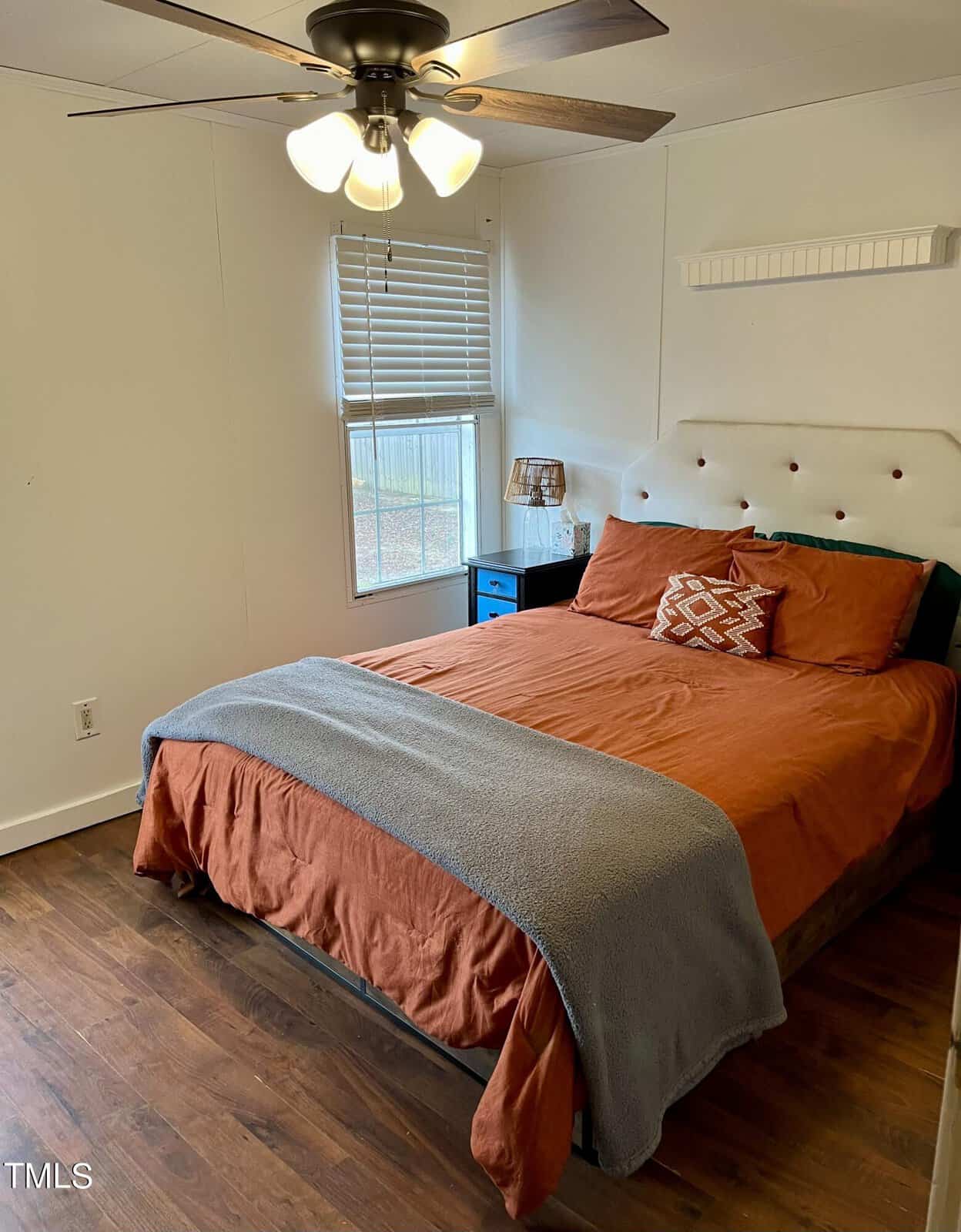 A cozy bedroom with a bed featuring orange bedding, a gray blanket, and patterned throw pillows. A small side table with a lamp is next to the bed, near a window with white blinds. The room has white walls, wooden floors, and a ceiling fan with lights.