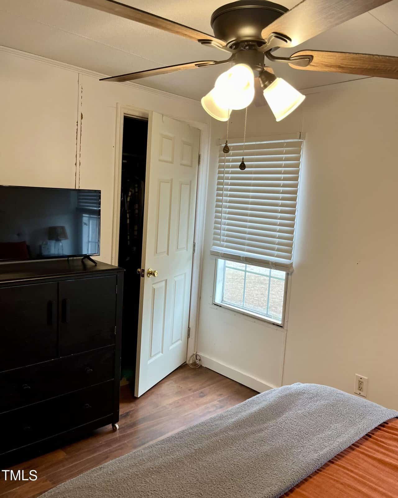 A cozy bedroom with white walls featuring a ceiling fan with lights. The room has a window with closed blinds and a small TV on a black dresser. An open closet door reveals some hanging clothes. A bed with an orange and gray blanket is partially visible.