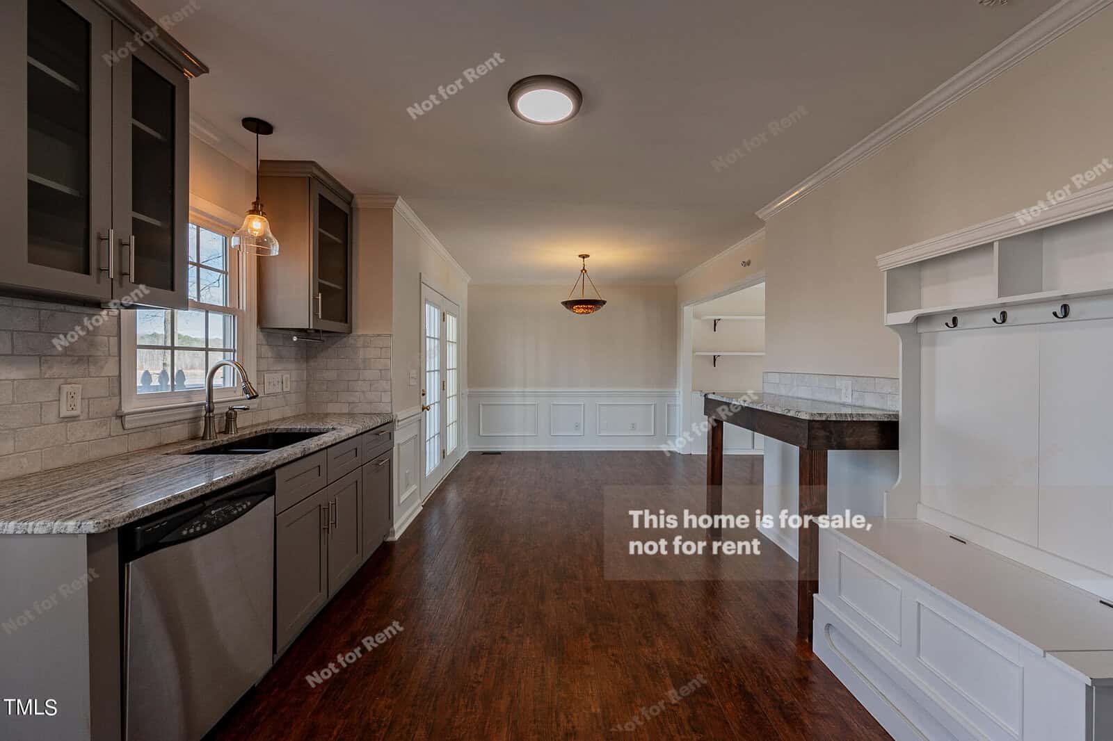 A modern kitchen with dark cabinets, granite countertops, and a stainless steel sink. It leads into a spacious dining area with dark wood flooring. Above, there's a round ceiling light, and the walls have wainscoting. The space is bright with natural light.