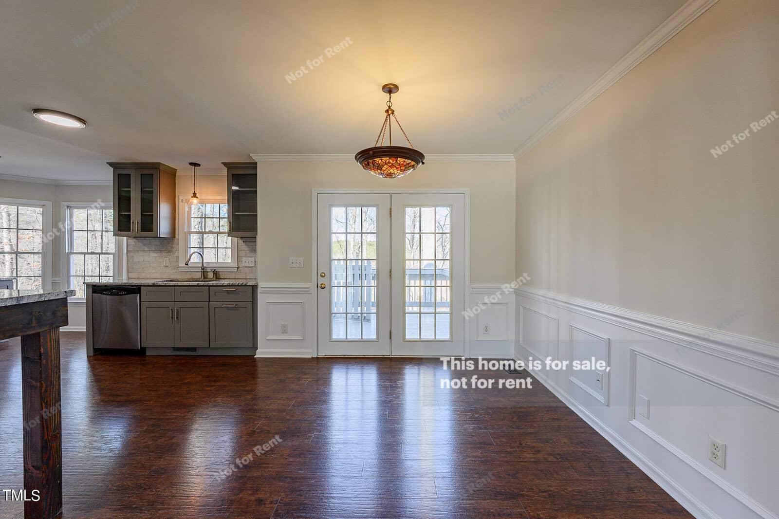 A spacious, empty kitchen and dining area with hardwood floors. It features a stylish chandelier, large windows with French doors opening to a deck, modern stainless steel appliances, and light-colored cabinets above a tiled backsplash.