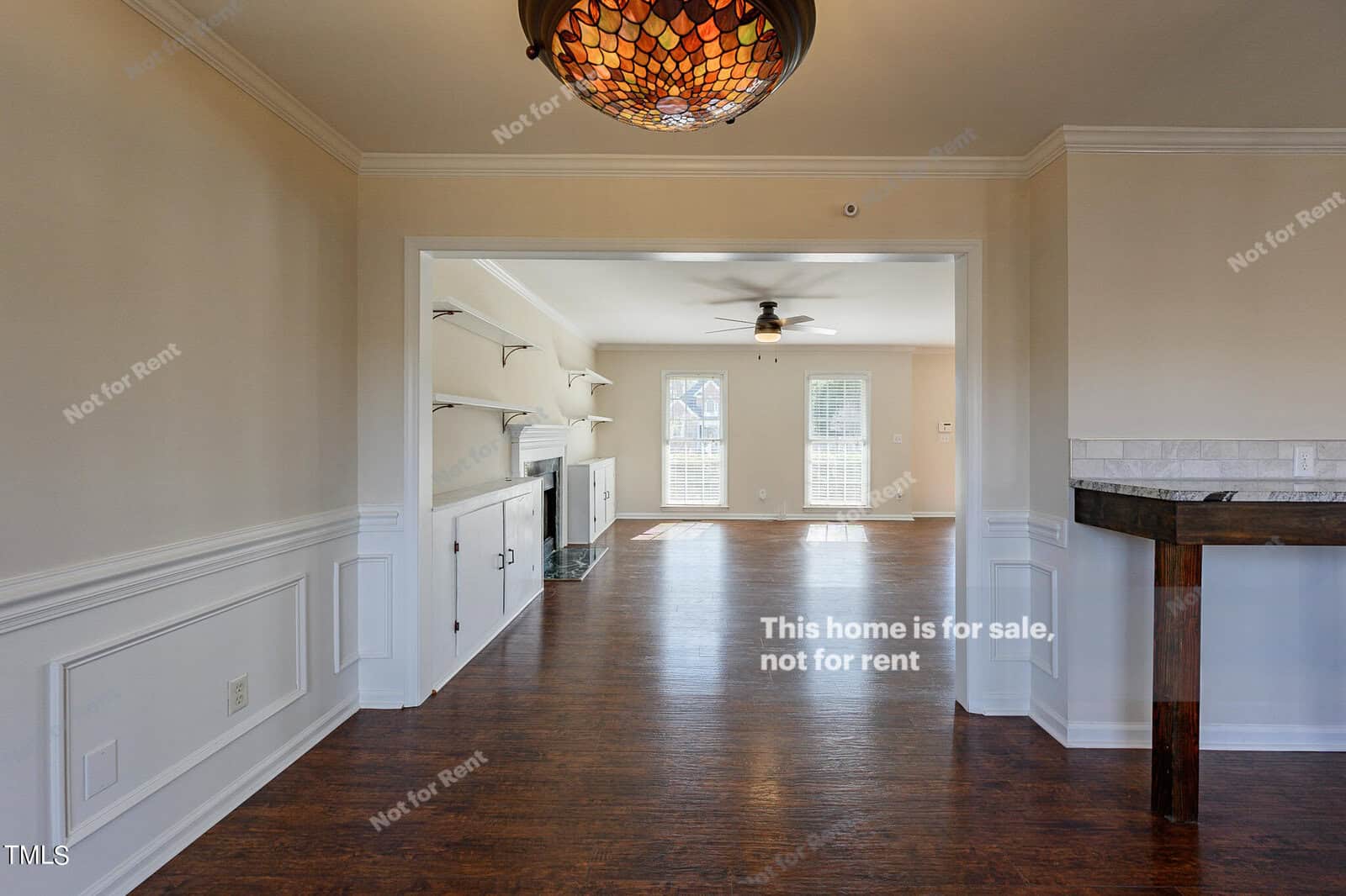 Interior of an unfurnished home with hardwood floors, neutral walls, and a decorative ceiling light. The space features a visible living area with built-in shelves and a large window, along with text reading, "This home is for sale, not for rent.