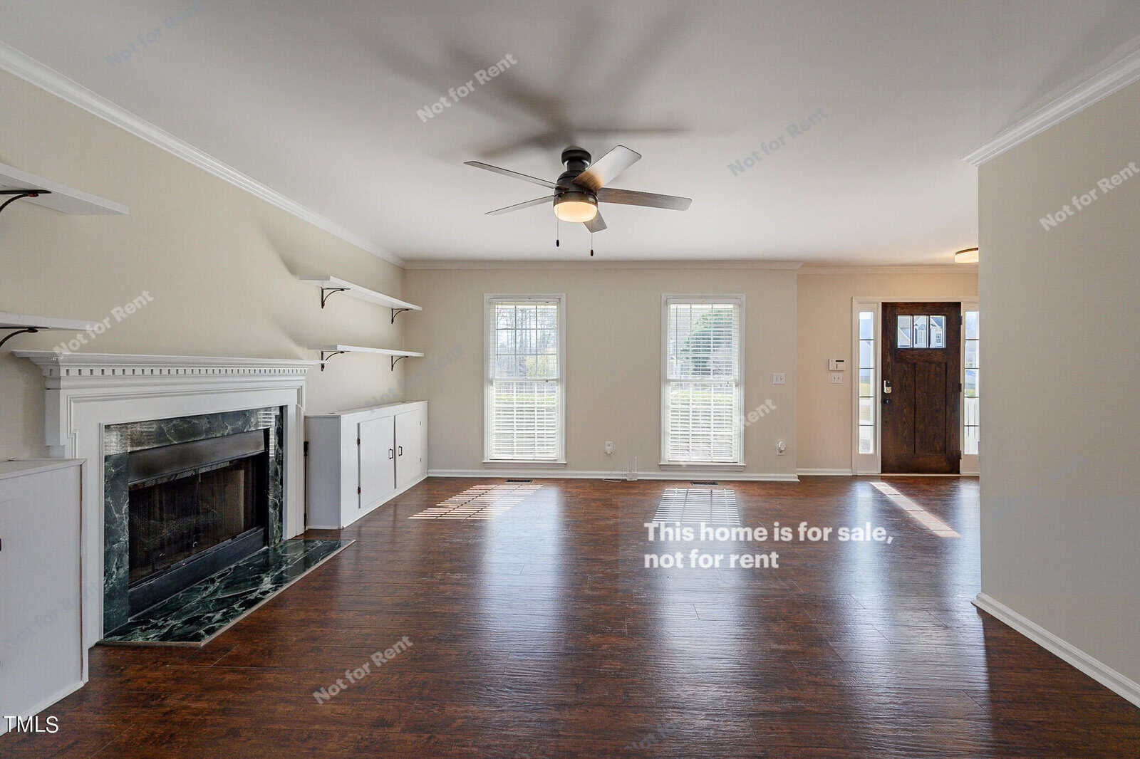 Spacious living room with hardwood floors, a fireplace, and shelves flanking each side. A ceiling fan is mounted above. Three large windows bring in ample light. The entrance is visible to the right with a wooden door. The walls are a light cream color.