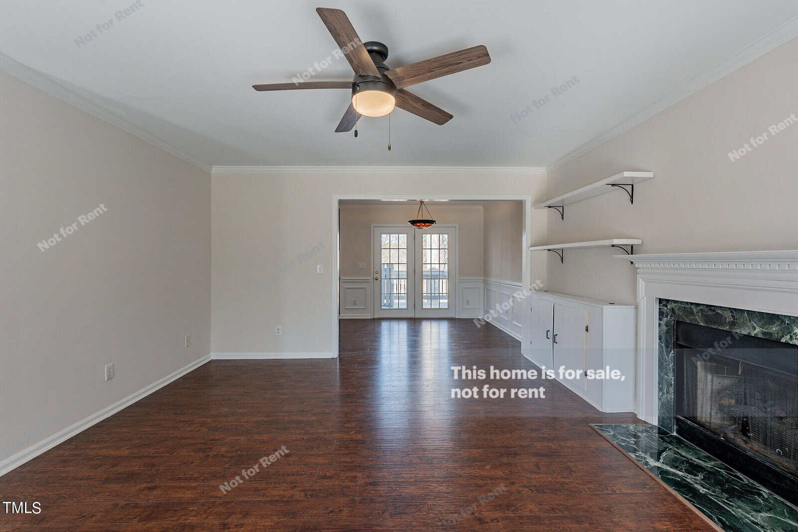 Spacious empty living room with hardwood flooring, a ceiling fan, and a white fireplace. Built-in shelves are next to the fireplace. French doors open to another room, and natural light fills the space. Walls have text indicating the home is for sale, not rent.