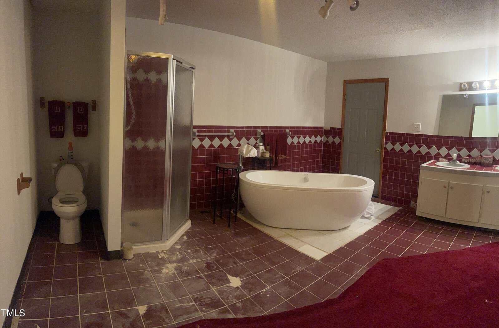 A bathroom with maroon and white tiles features a glass shower, a toilet, a white freestanding bathtub, and a sink with a mirror. Red accents are present, including carpet and towels on the wall.