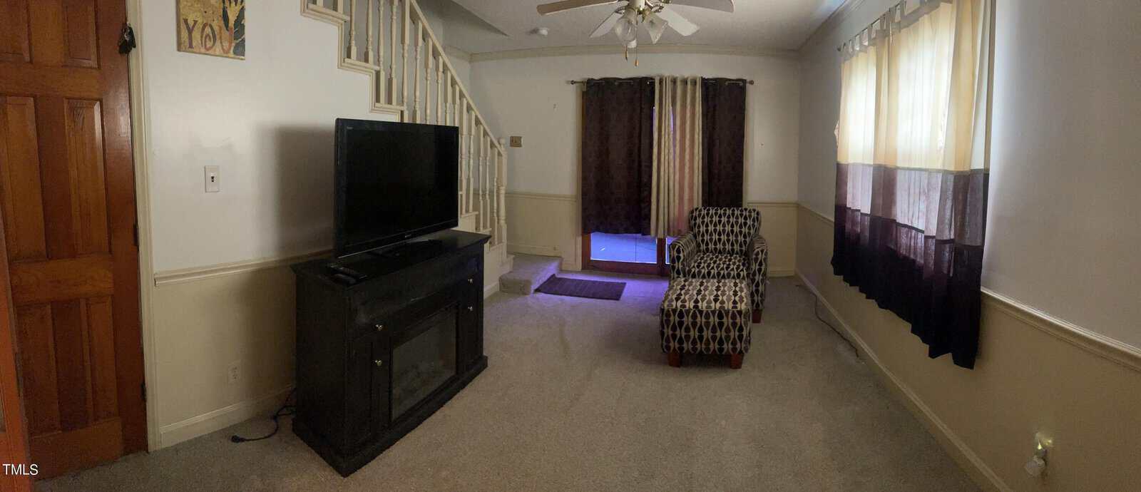 A cozy living room with beige walls and carpet, featuring a patterned armchair and a flat-screen TV on a dark wooden stand. A staircase leads up on the left, and large windows with sheer and dark curtains allow natural light in.