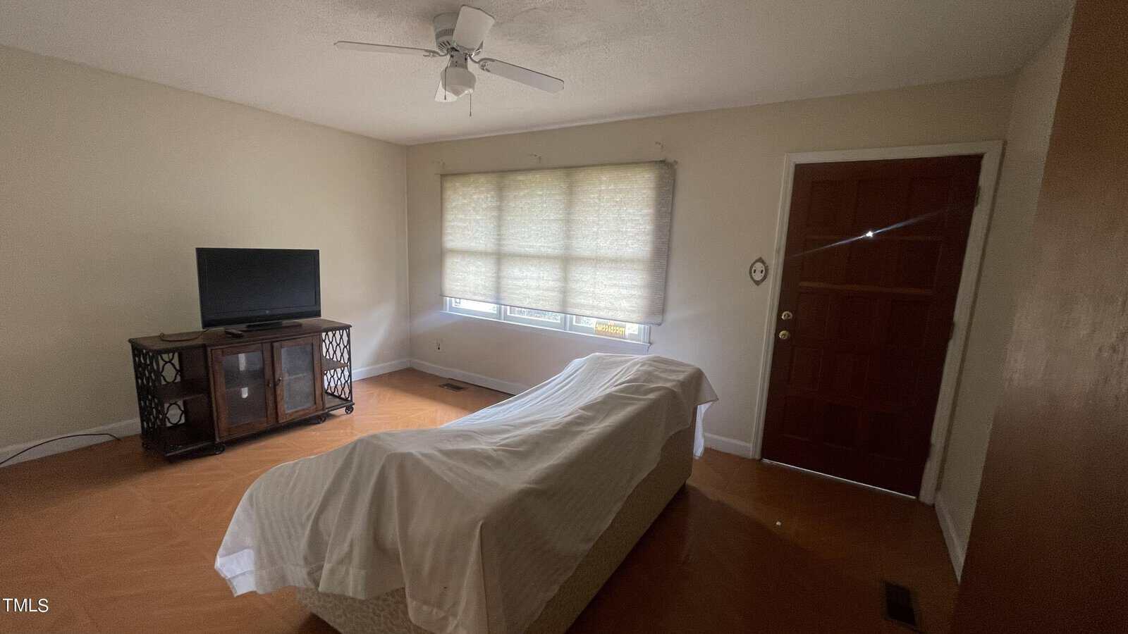 A living room with a large window covered by blinds, a ceiling fan, a wooden door, a TV on a stand, and a couch covered with a white sheet. The room has wooden flooring and neutral-colored walls.