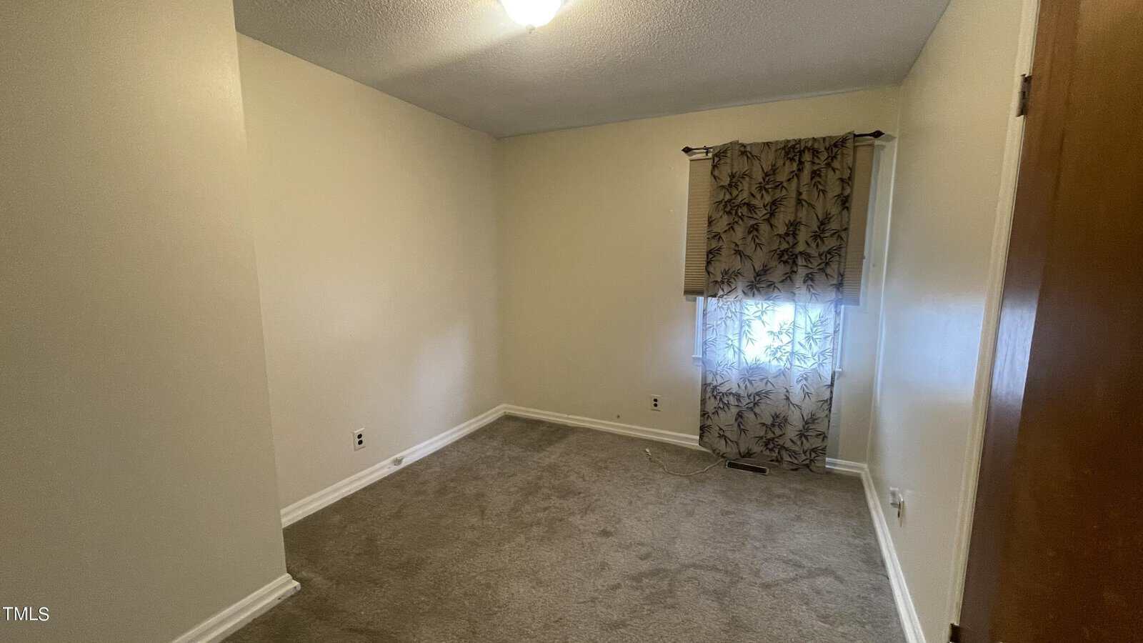 A small, empty room with beige walls and gray carpeting. A single window is covered with a decorative sheer curtain featuring a leaf pattern. The room has a ceiling light and baseboard molding.