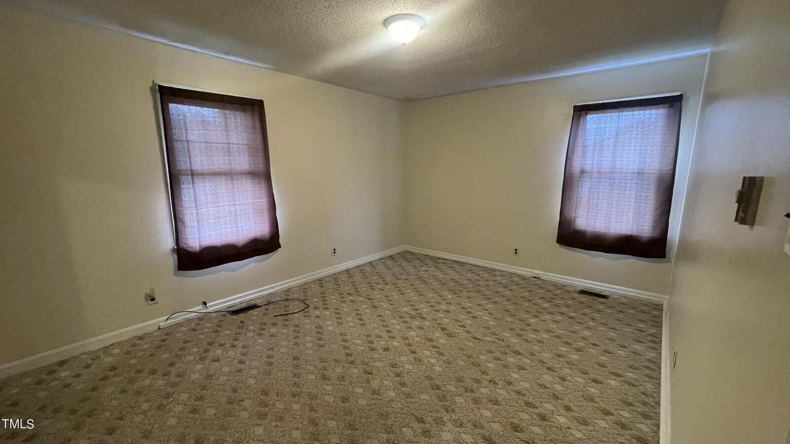 A vacant room with beige carpet and light-colored walls. Two windows with brown curtains allow soft light inside. A ceiling light fixture is centered above. A cable can be seen lying on the floor.