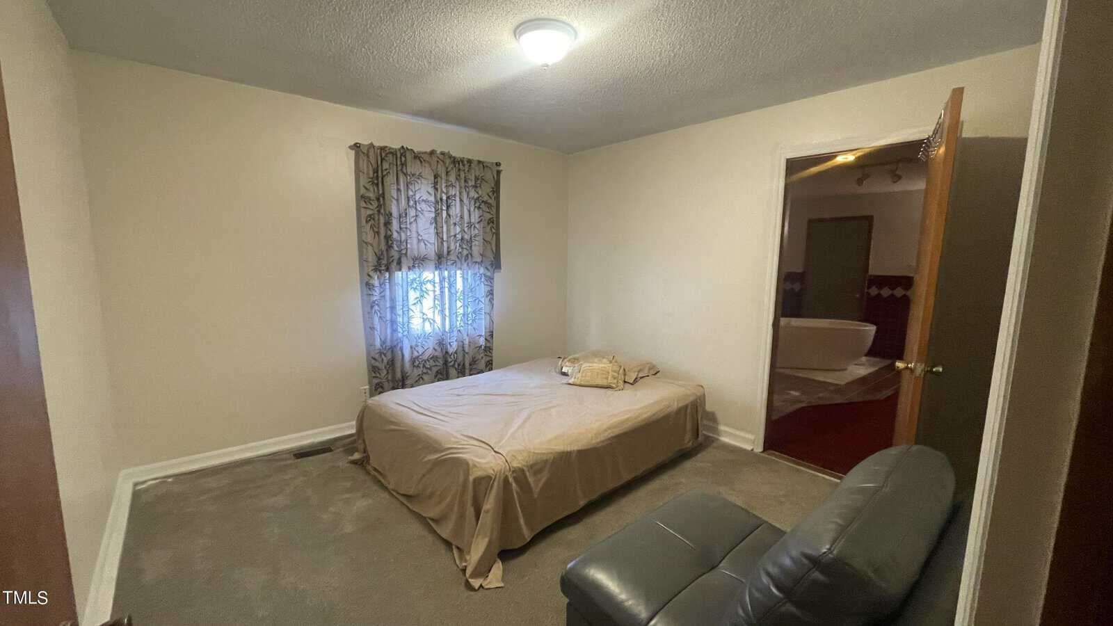 Small, carpeted bedroom with beige walls and a single bed covered in a beige blanket. A window with patterned curtains is on the left. There is a leather chair and an open door leading to a bathroom. The room is lit by a ceiling light.