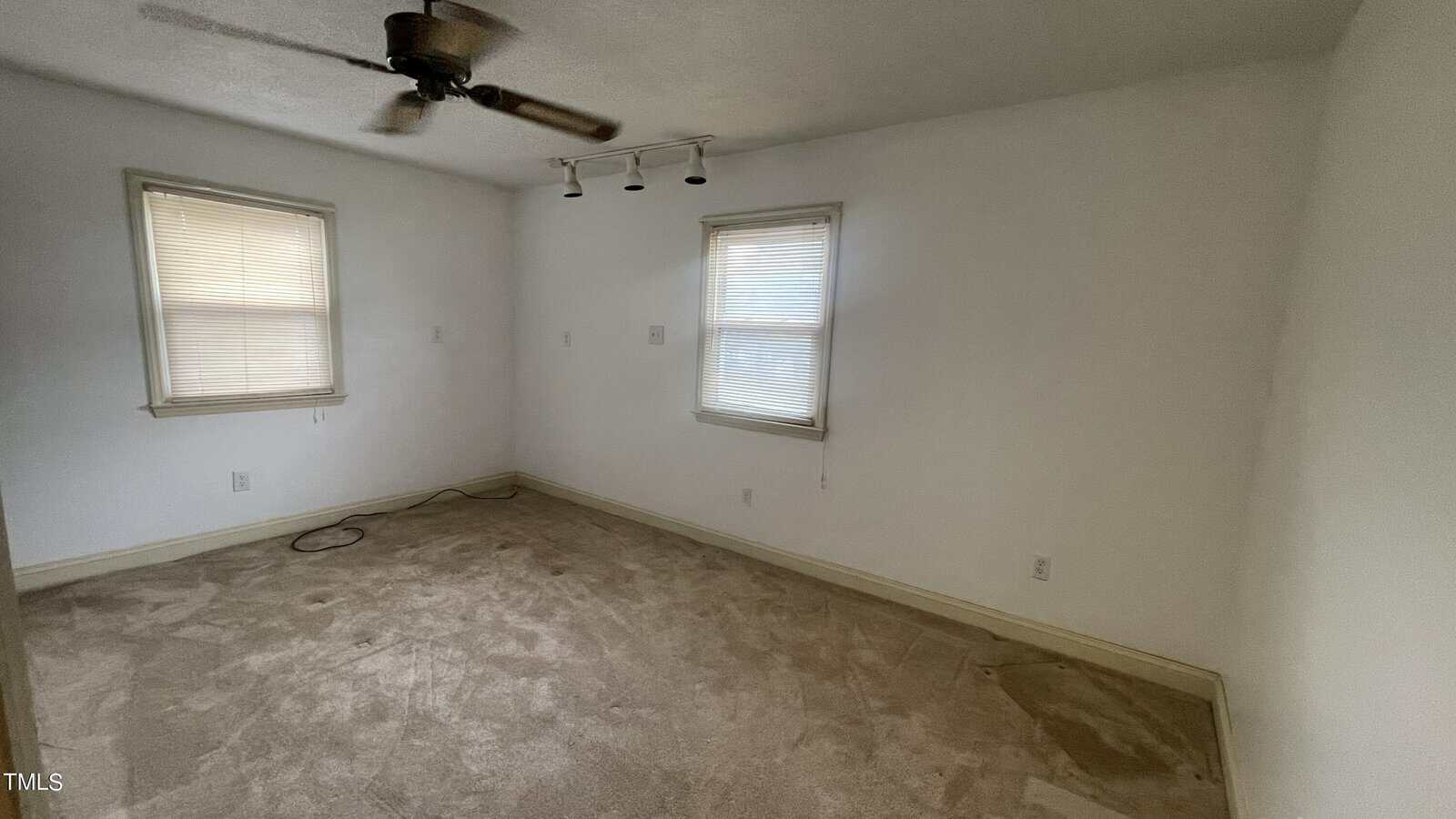 An empty room with two windows covered by blinds, beige carpet, and a ceiling fan. A wall features track lighting, and a few electrical cords lie on the floor. Walls are painted white. Natural light enters through the windows.