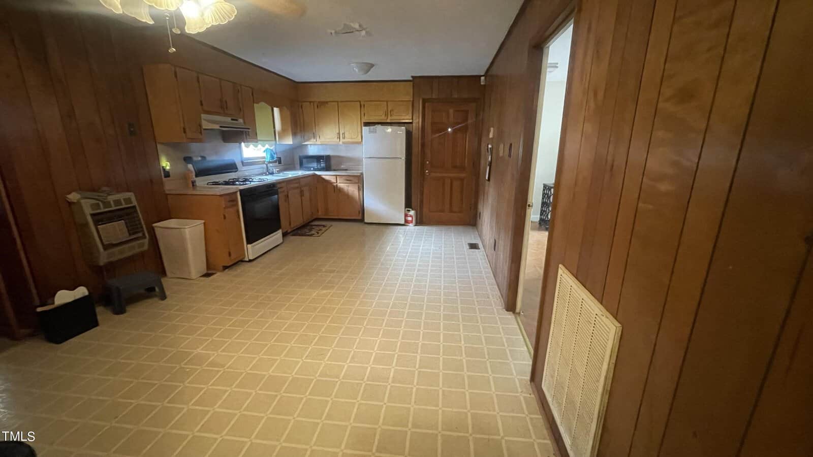 A kitchen with wooden cabinets, a white refrigerator, stove, and oven. The floor is covered with light-colored linoleum. A ceiling fan is visible at the top and a doorway leads to another room on the right. Wood-paneled walls surround the space.