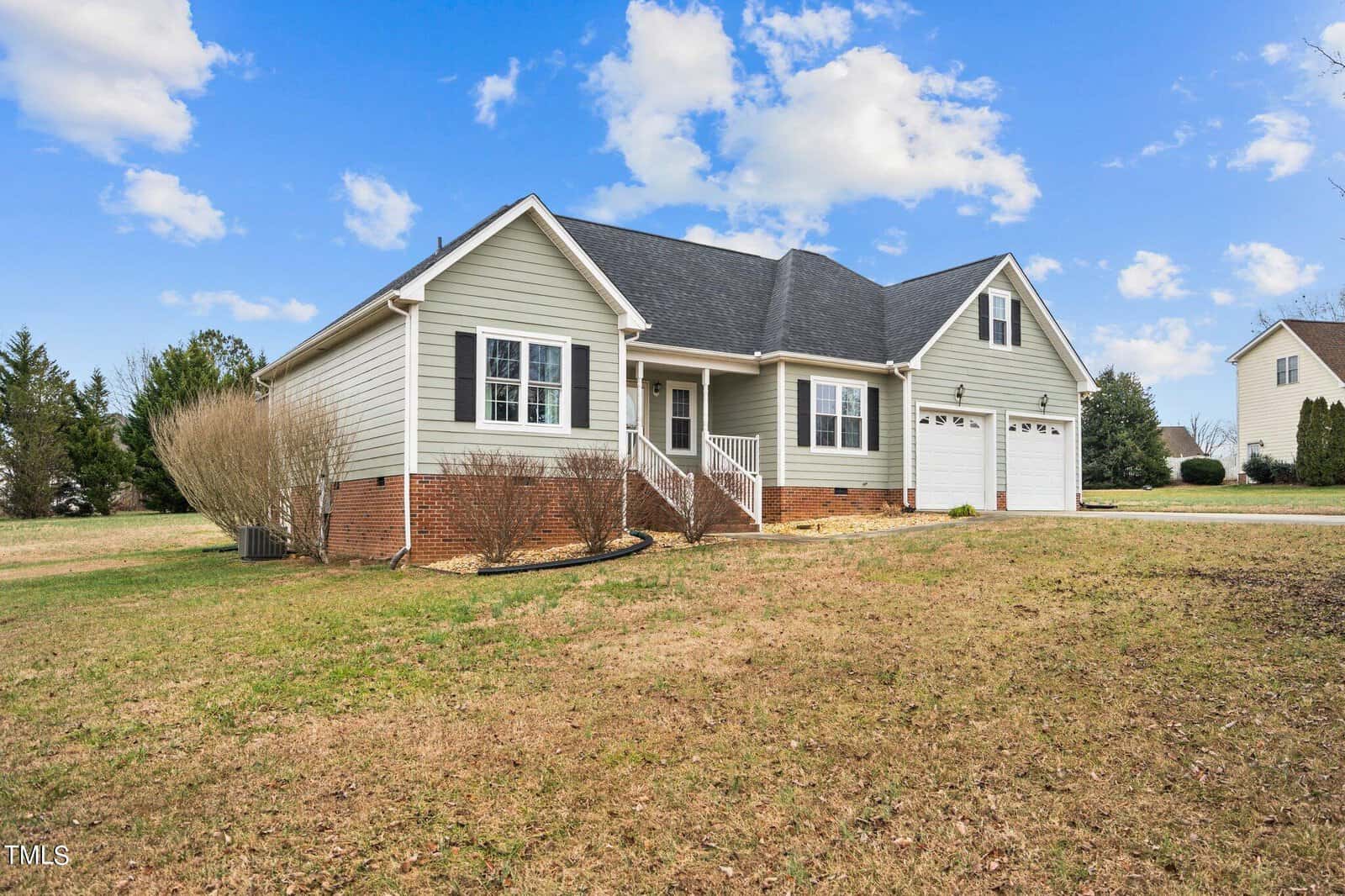 A single-story house with light gray siding, white trim, and a dark roof. It has a front porch, two garage doors on the right, and a landscaped yard with sparse grass and bushes. The sky is partly cloudy.