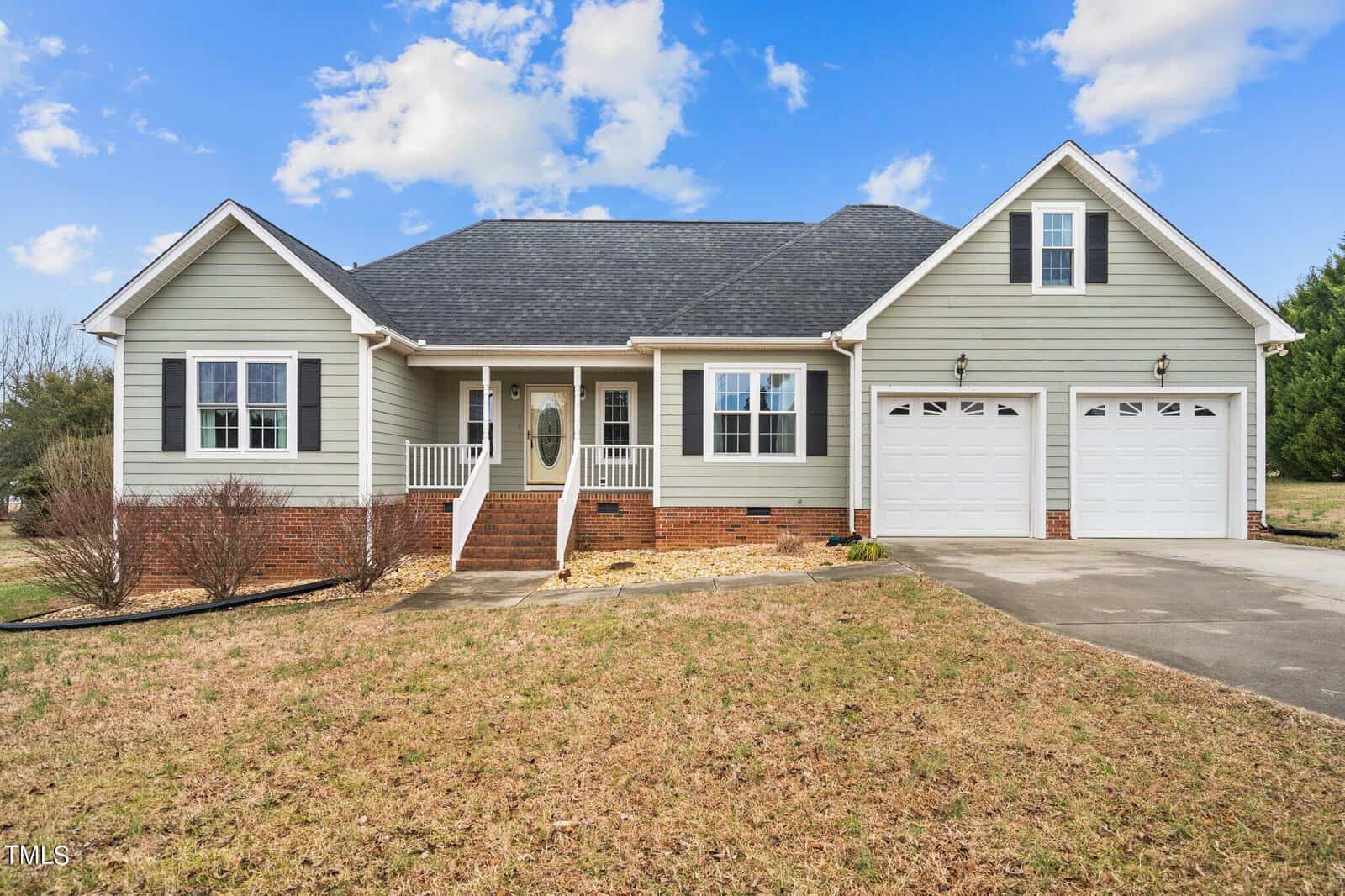 A charming single-story house on 1066 Wheeler Pond Road in Creedmoor, boasting a gray facade, white trim, and dark roof. It features a front porch with white railings and two garage doors, with a pathway leading to the entrance amidst a grassy yard under a blue sky with scattered clouds.