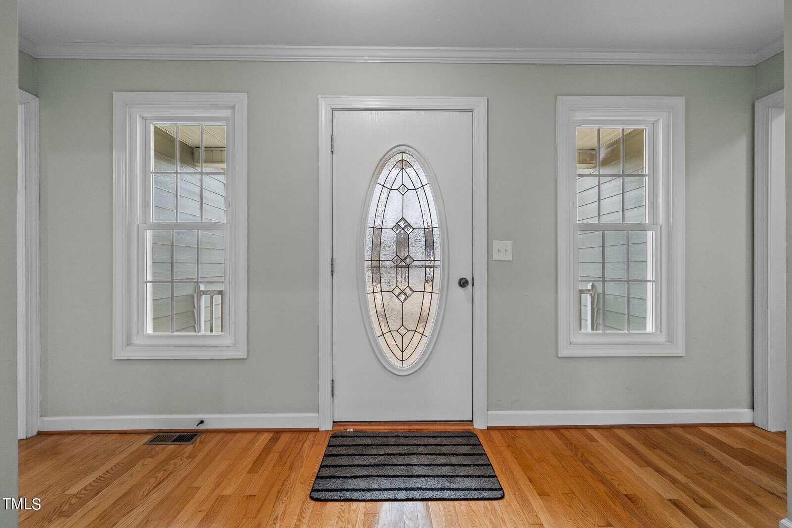 A white oval glass-paneled front door flanked by two rectangular windows. The walls are light green, and the hardwood floor is a warm brown. A black striped doormat is placed in front of the door.