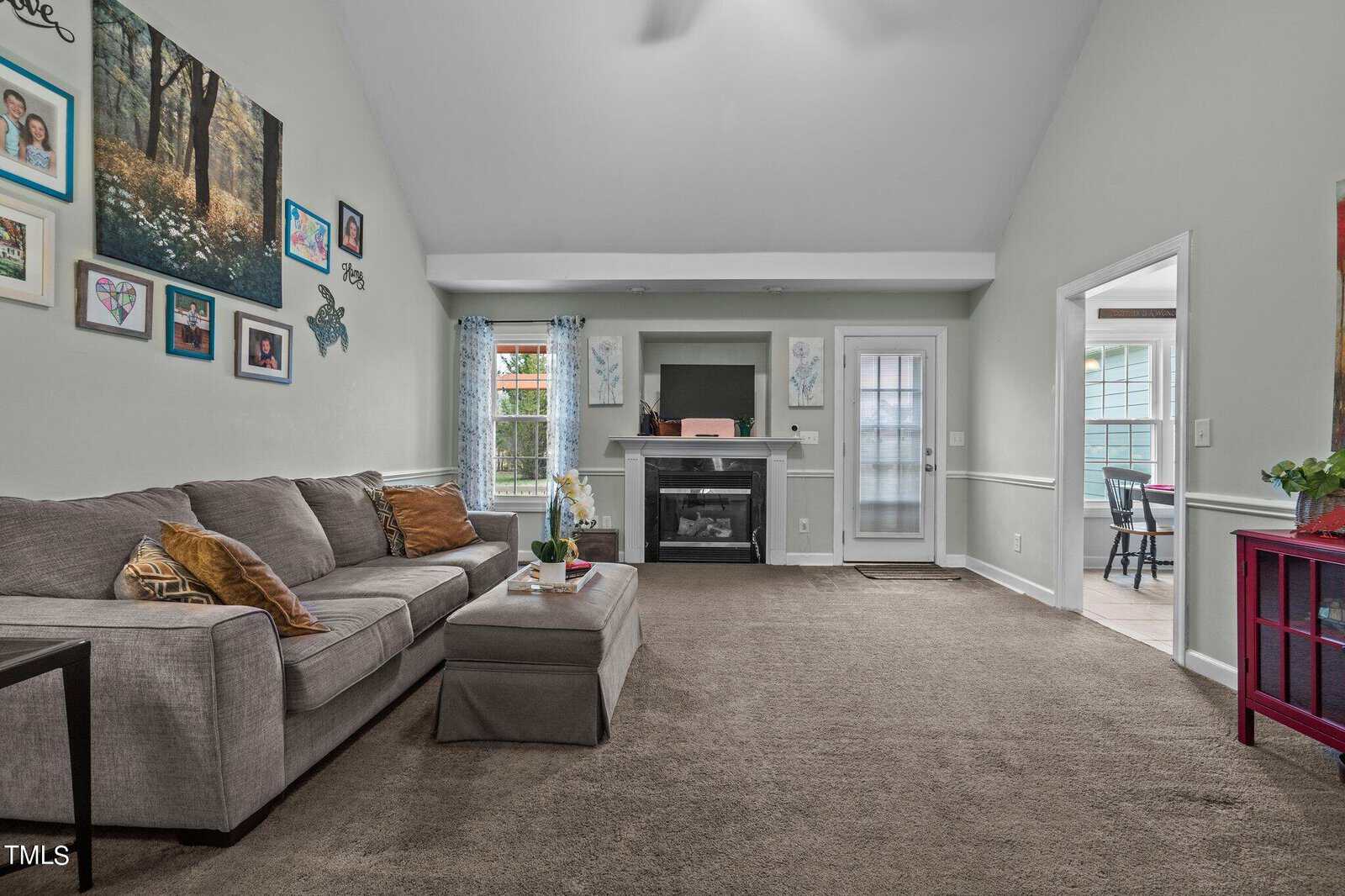 A spacious living room with a vaulted ceiling. It features a gray sectional sofa, a fireplace with a TV above, and a wall displaying family photos. A door leads to an outdoor area, and there are large windows with blue curtains letting in natural light.