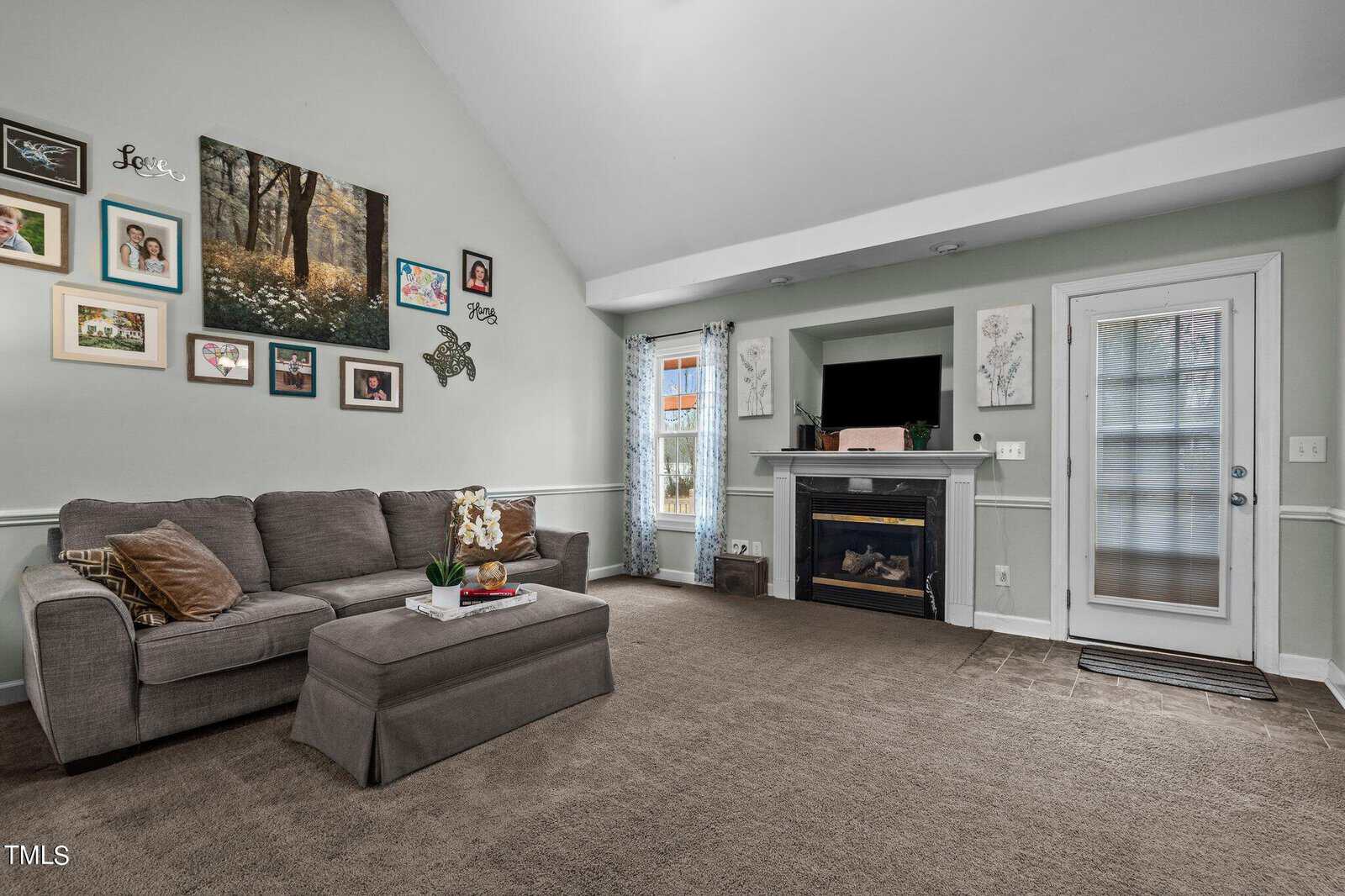 A cozy living room with a green sectional sofa and ottoman, decorated wall with family photos and artwork, a fireplace with a TV above it, and a door with a curtain. The carpet is brown, and natural light fills the room.