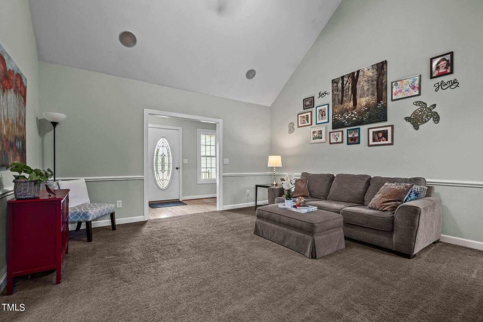 A cozy living room with a high ceiling, featuring a gray sectional sofa and coffee table. The walls are adorned with various photo frames and artwork. The room is well-lit by a floor lamp beside the sofa, and a doorway leads to a sunlit area.