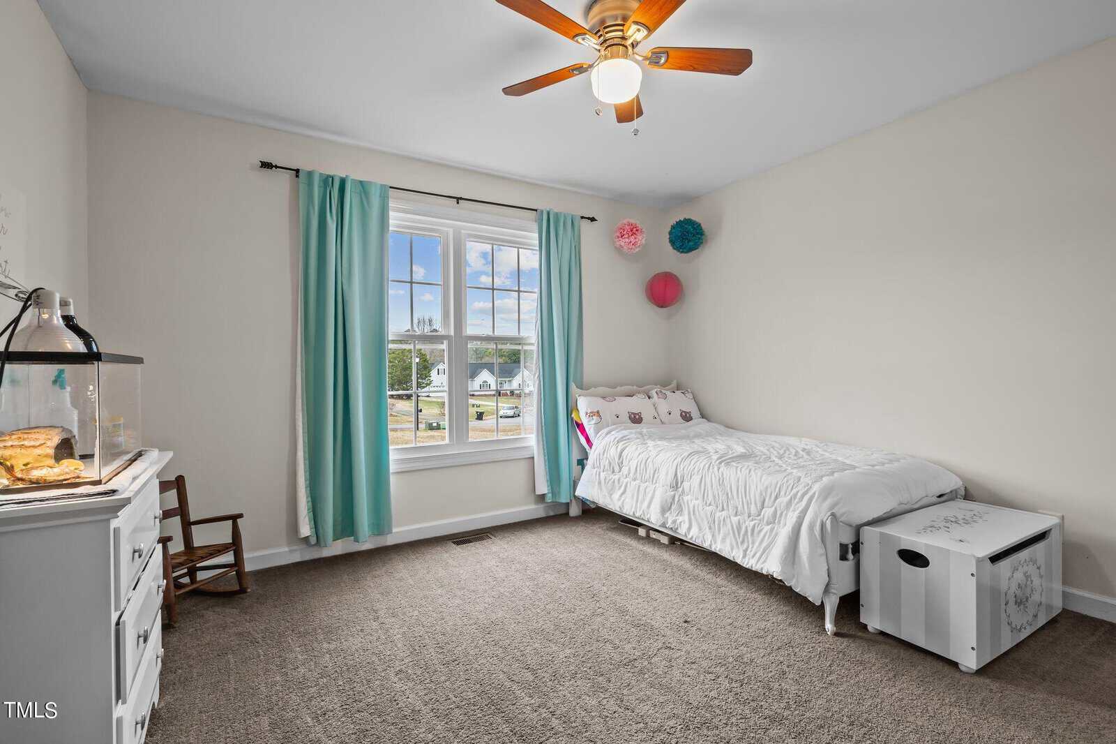 Bedroom with a single bed against the wall, decorated with colorful hanging ornaments. A window with teal curtains lets in natural light. A white dresser holds a fish tank, and a small rocking chair is near the corner. The room has beige carpet.