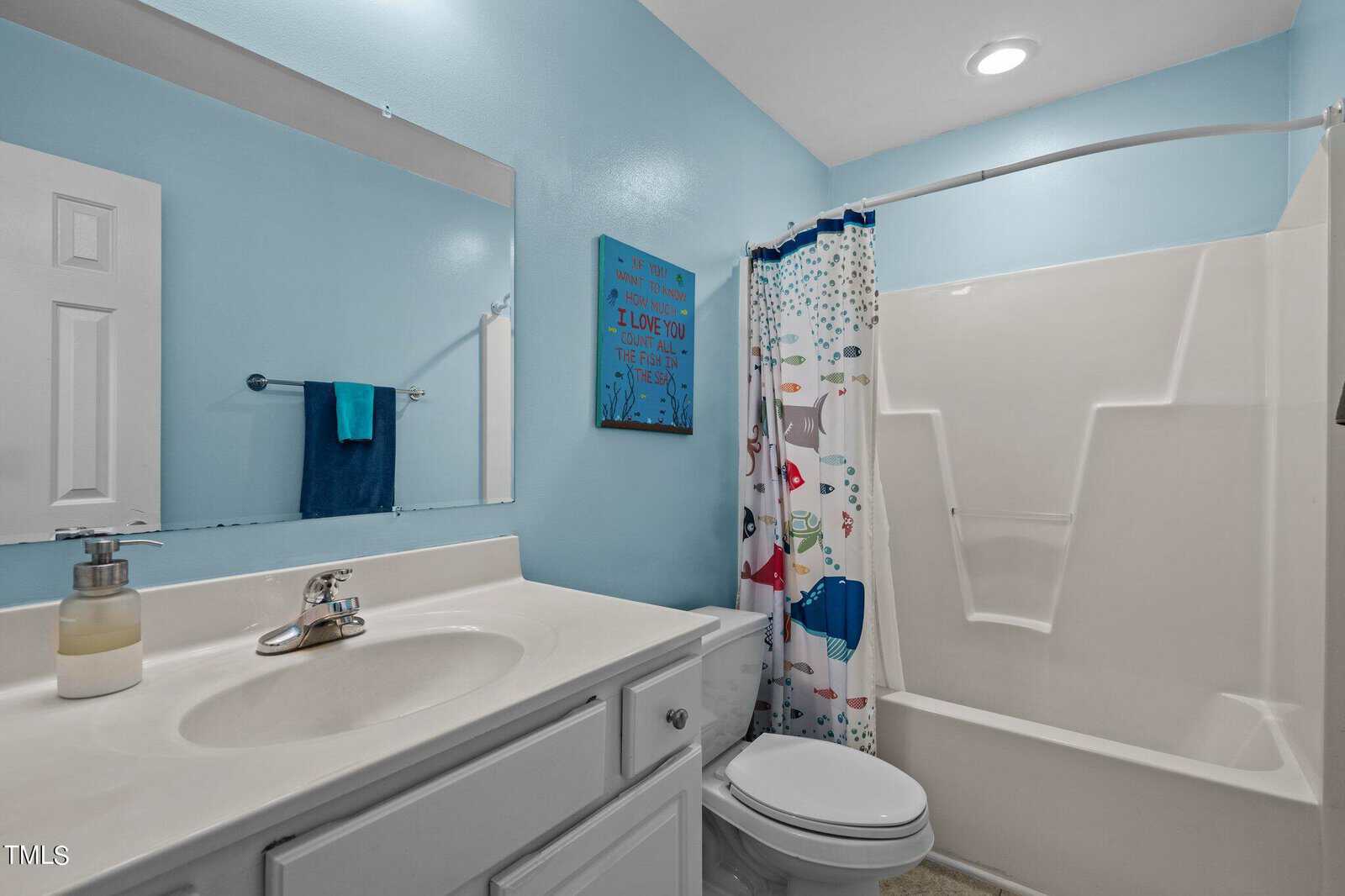 A clean bathroom with light blue walls features a white vanity with a sink and a mounted mirror above. A decorative picture hangs on the wall. A shower with a colorful curtain is next to a toilet.