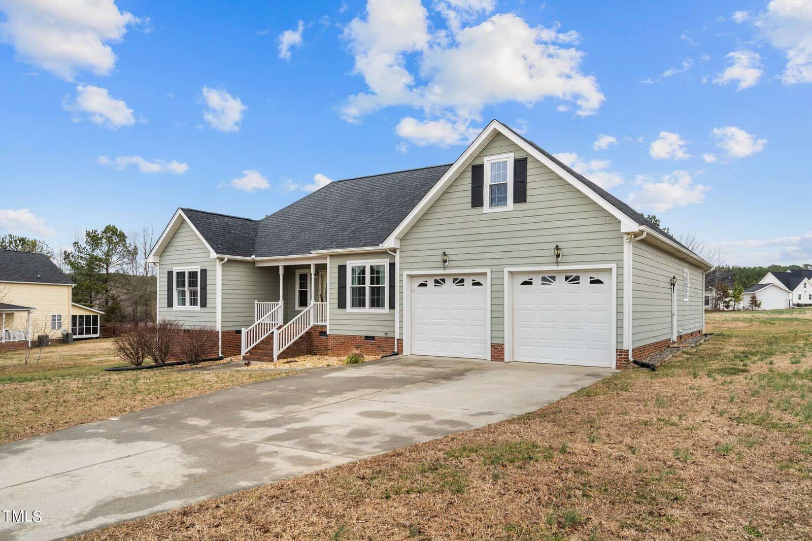 A single-story house with light gray siding, two white garage doors, and a dark gray roof. It has a small front porch with white railings. The house sits on a dry, grassy yard under a blue sky with scattered clouds.