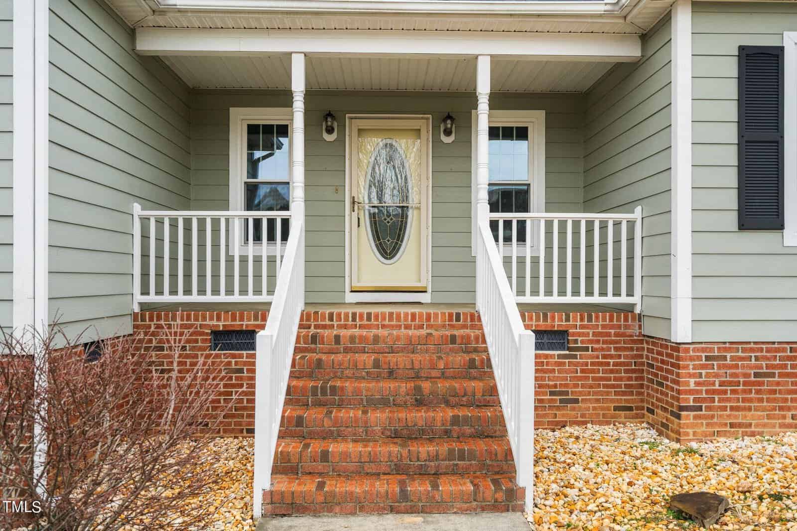 The image shows the front entrance of a house with green siding. It features a brick staircase leading to a wooden door with an oval glass panel. White railings and two windows flank the door. Sparse landscaping with stones and bare bushes is visible.