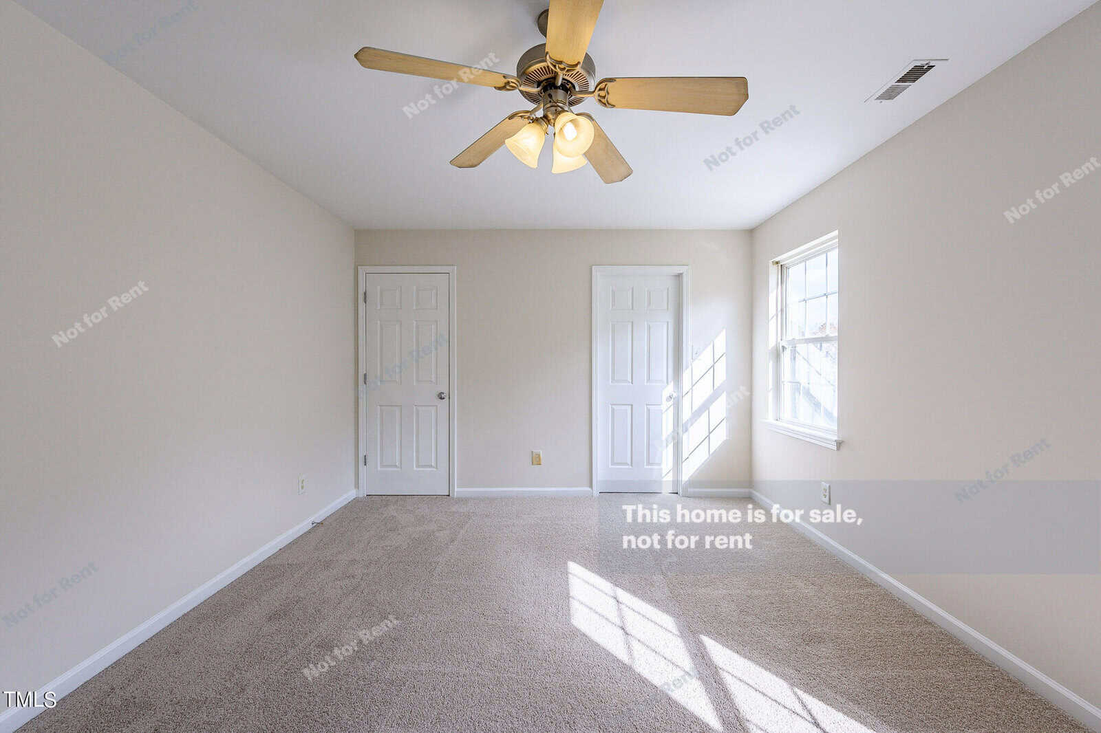 An empty carpeted room with beige walls, a ceiling fan with lights, two closed white doors, and a window letting in natural light. The wall reads, "This home is for sale, not for rent.