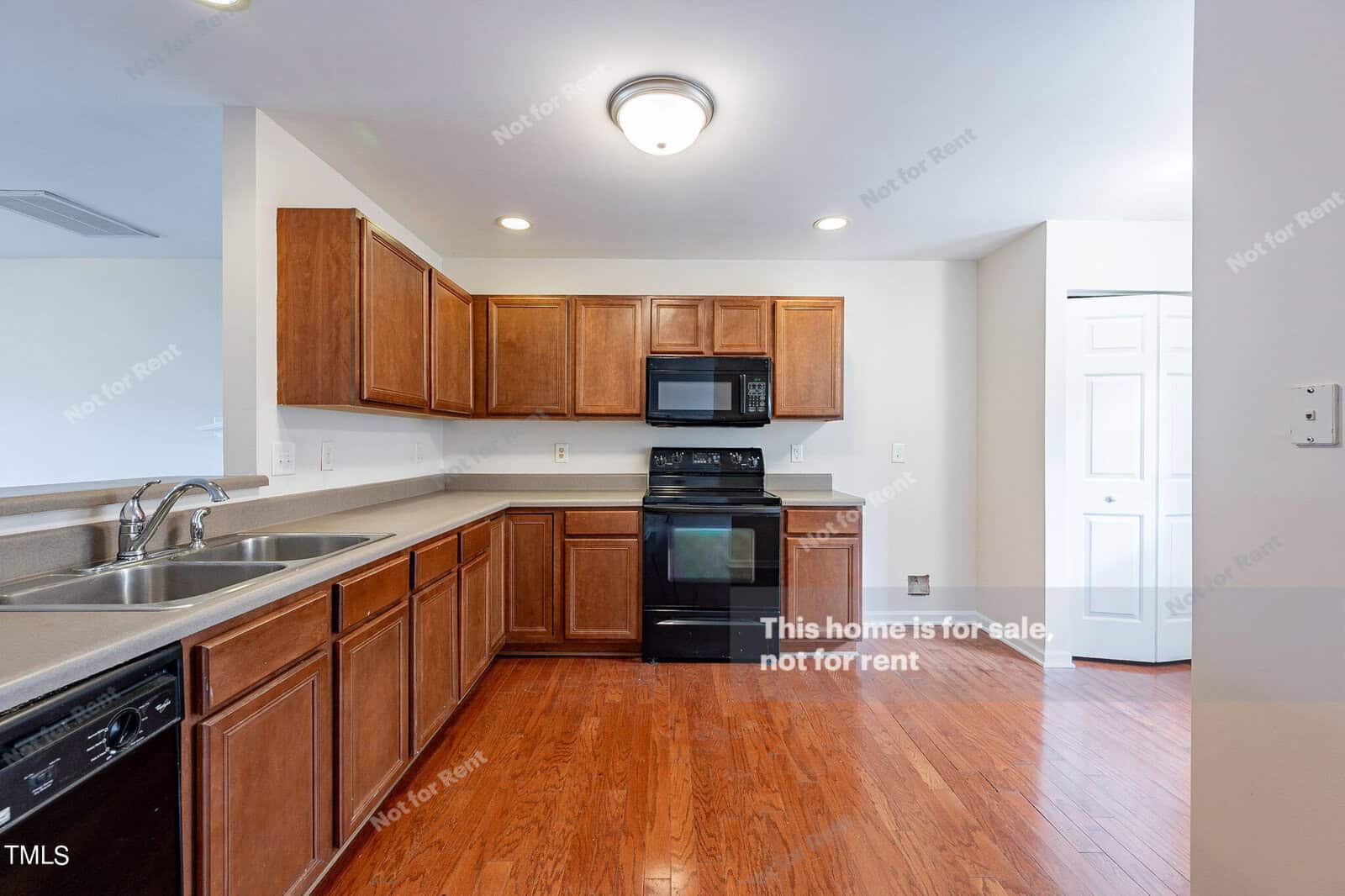 A modern kitchen with wooden cabinets, black appliances including an oven and dishwasher, and a double sink. The space features hardwood floors and bright lighting. Walls have "Not for Rent" notices.