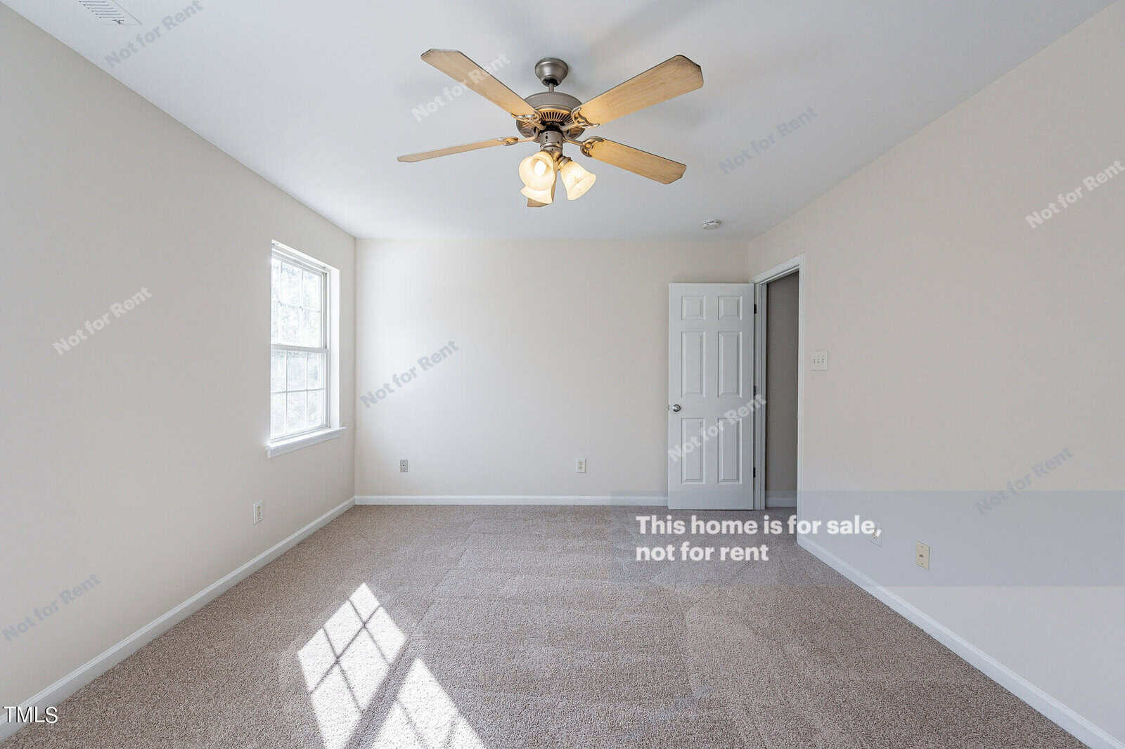 Bright, empty room with beige carpet, cream walls, and a ceiling fan with lights. A large window lets in natural light, and a white door is visible on the right. Text overlay indicates the home is not for rent.