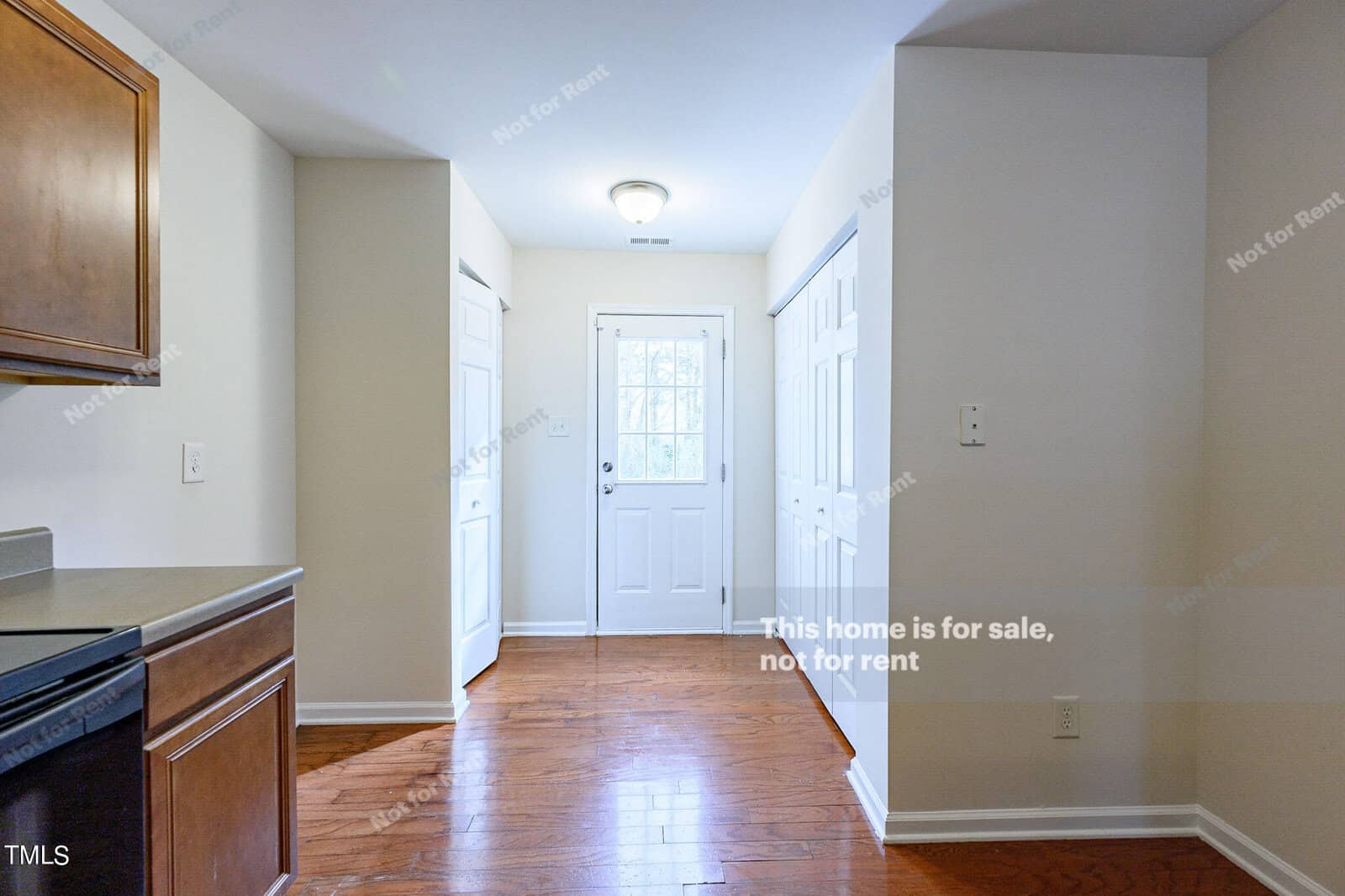 A bright, empty room with wooden floors and white walls. There are two white doors, one with a window, with "This home is for sale, not for rent" written on the image. A partial view of a kitchen counter is visible on the left.
