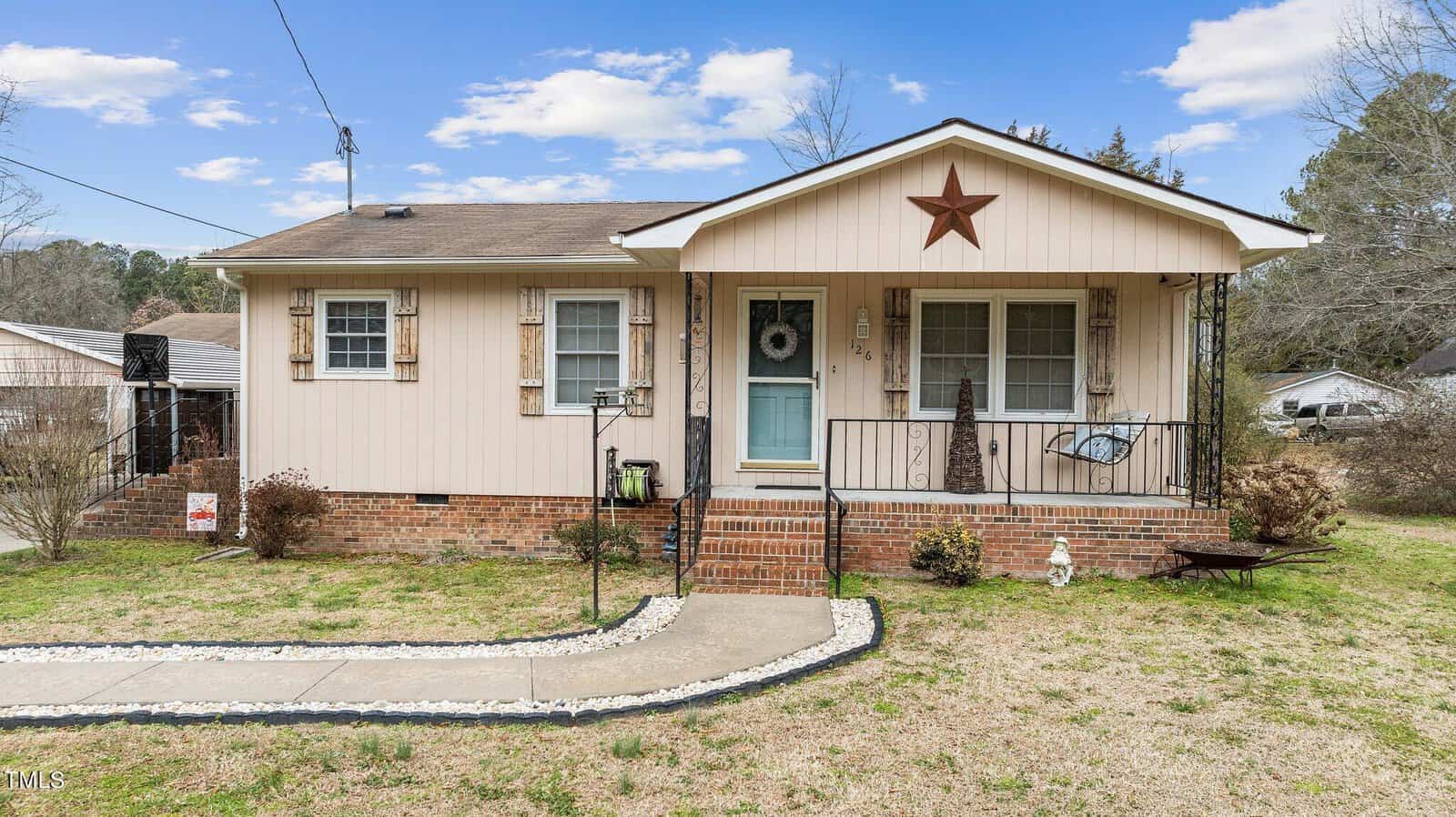 A small single-story house at 126 S Elizabeth Street, Henderson NC, with beige siding and a brick foundation, featuring one red star decoration above the porch and a neatly maintained lawn.