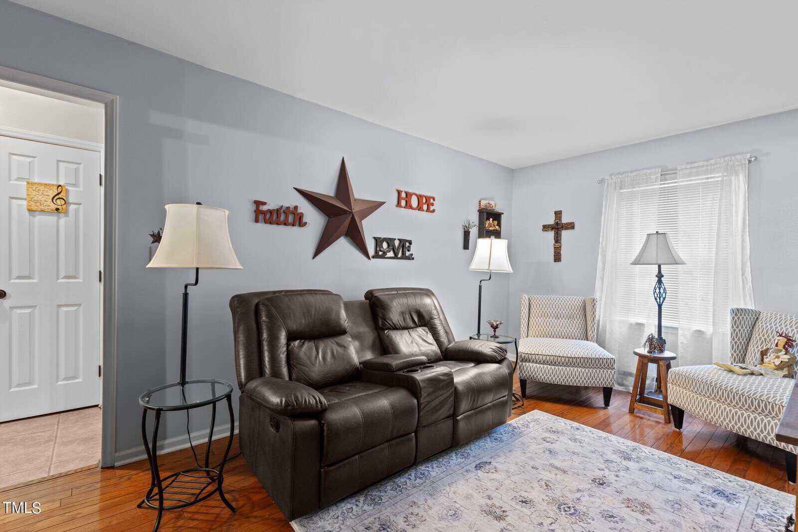 A neatly arranged living room featuring a brown leather sofa, two patterned armchairs, a white rug, and decorative wall hangings that include the words "faith," "hope," and "love.