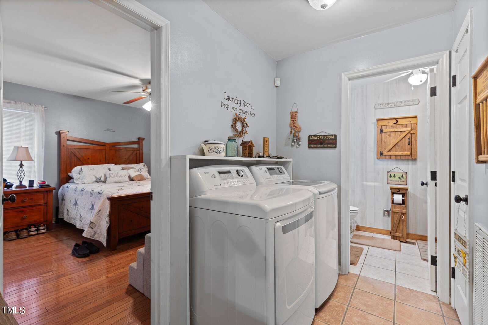 A laundry room with a washer and dryer is adjacent to a bedroom with a wooden bed frame and a nightstand; a bathroom is visible through an open door.