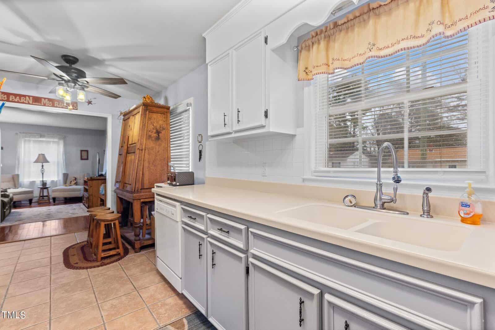 A well-lit kitchen area with white cabinetry and beige countertops, featuring a double basin sink and a dishwasher. the kitchen opens up to a living room area with hardwood floors and a ceiling fan.