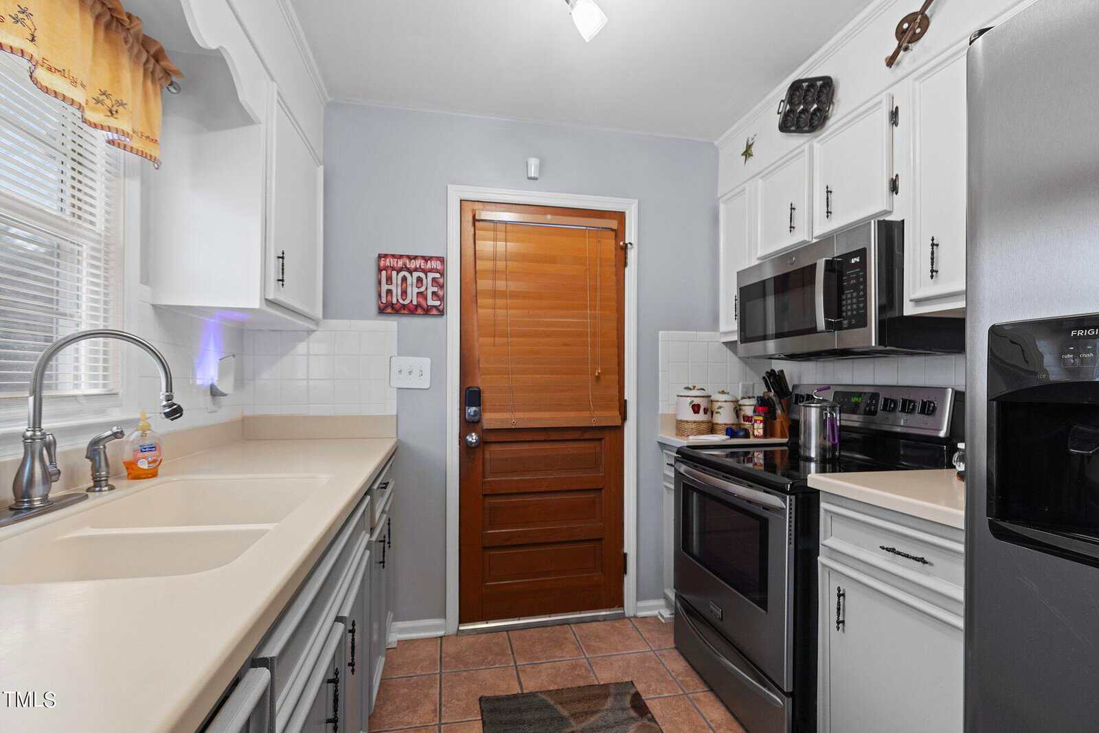 A modern kitchen with white cabinetry, black appliances, and a wooden door with a decorative "hope" sign hanging on it.