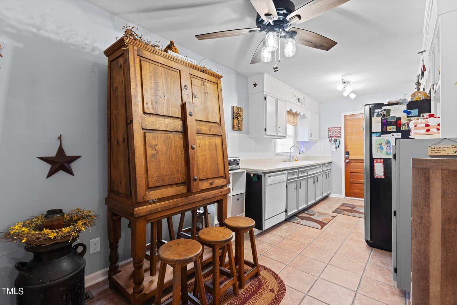 A neatly furnished kitchen with white cabinetry, a central wooden cabinet with a hutch, bar stools, and a ceiling fan, showing a mix of modern appliances and rustic decor elements.