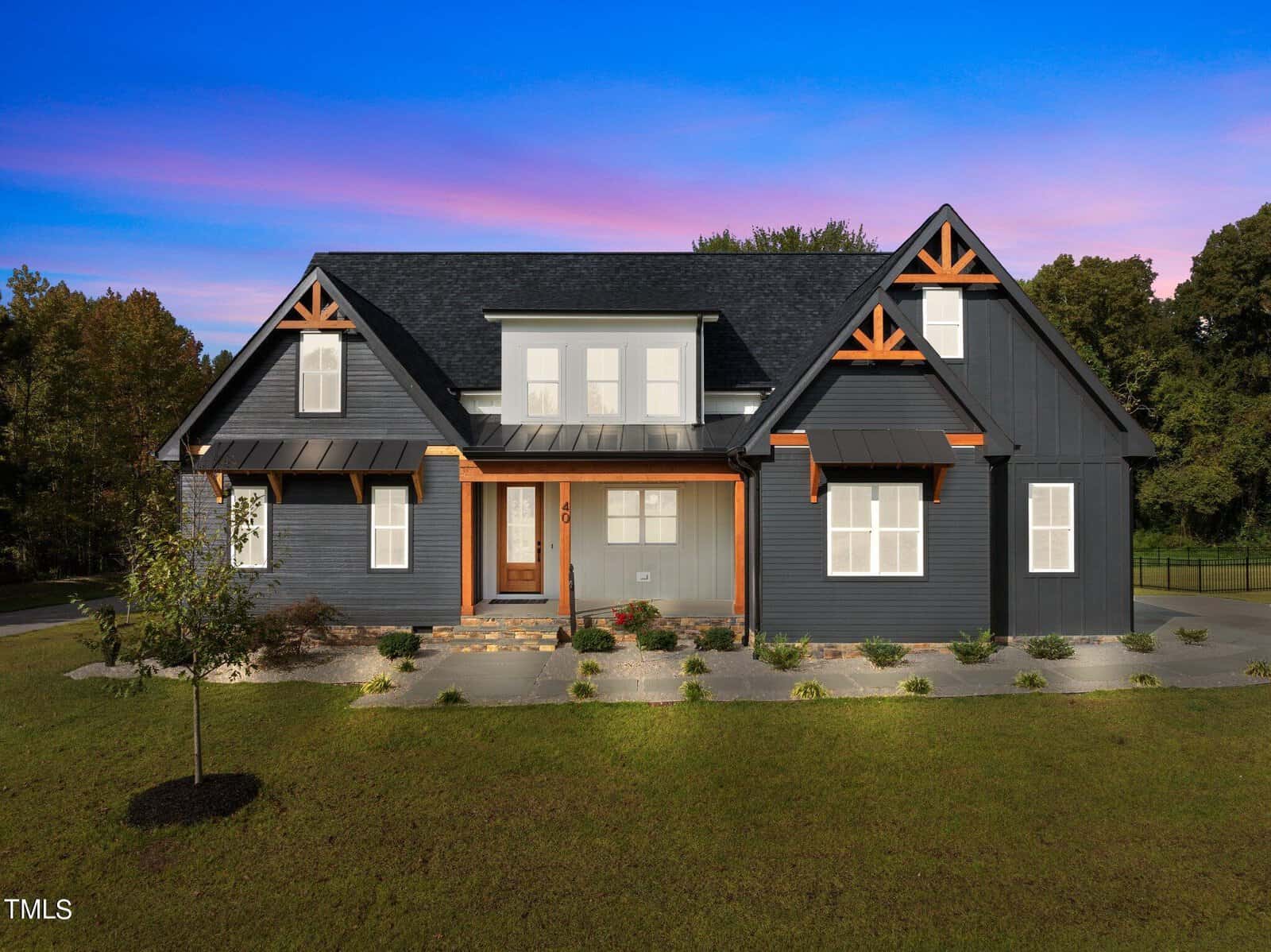 A modern, dark gray house with white trim and wooden accents sits on a large, well-kept lawn under a vibrant blue and purple sunset sky, surrounded by trees.