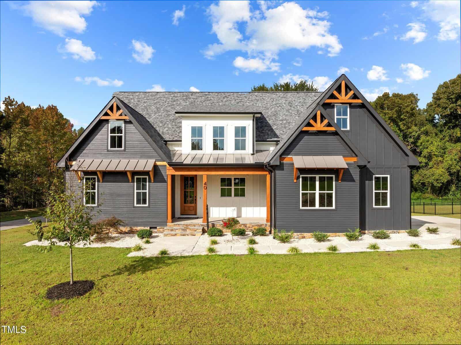 A modern farmhouse-style home with dark gray siding, white trim, wood accents, and a manicured lawn, set against a backdrop of trees and blue sky with scattered clouds.