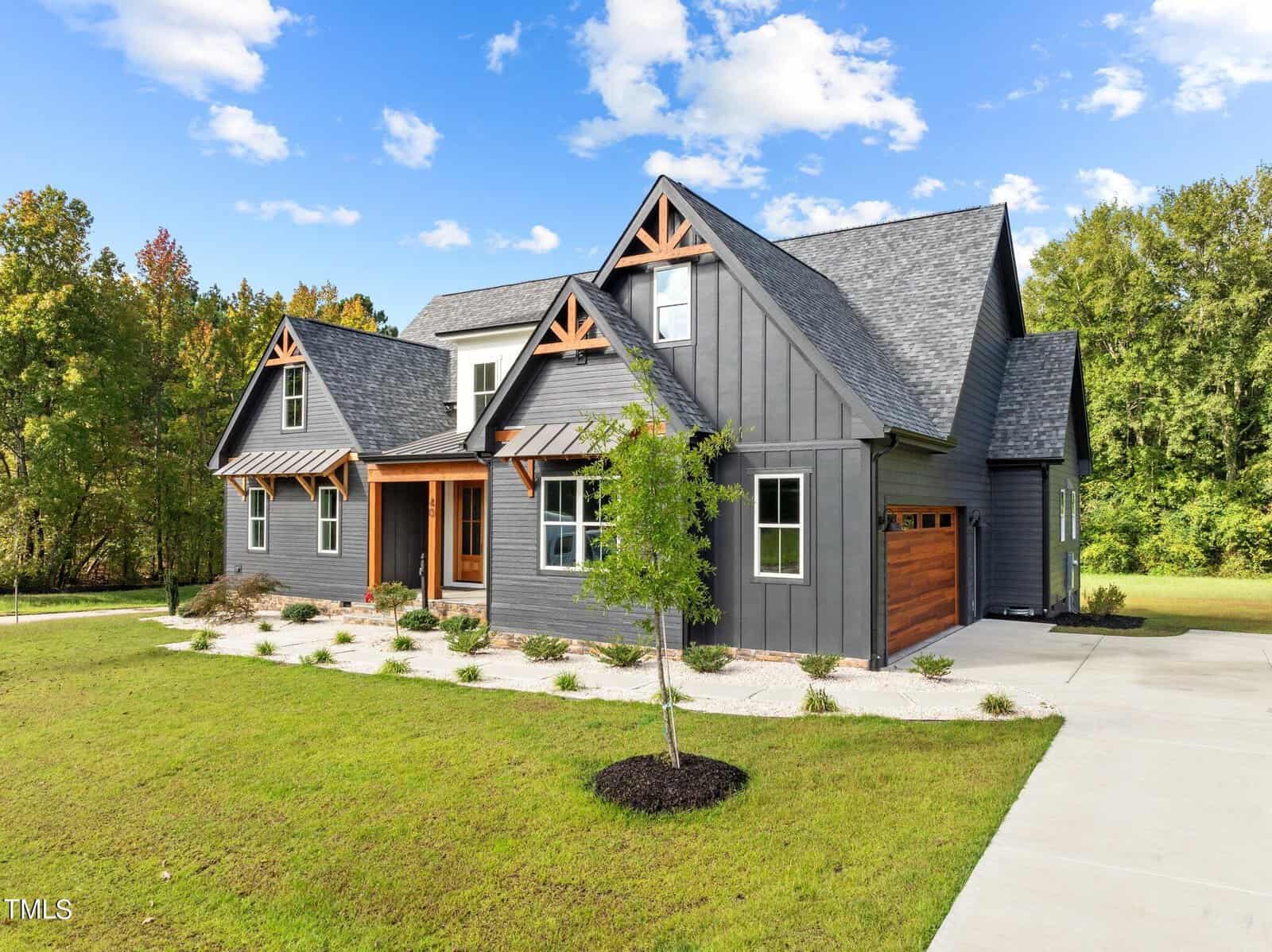Modern two-story house with dark gray exterior, wood accents, and a neatly landscaped front yard with a small tree and green grass. The home features a gabled roof and a double garage on the right side.