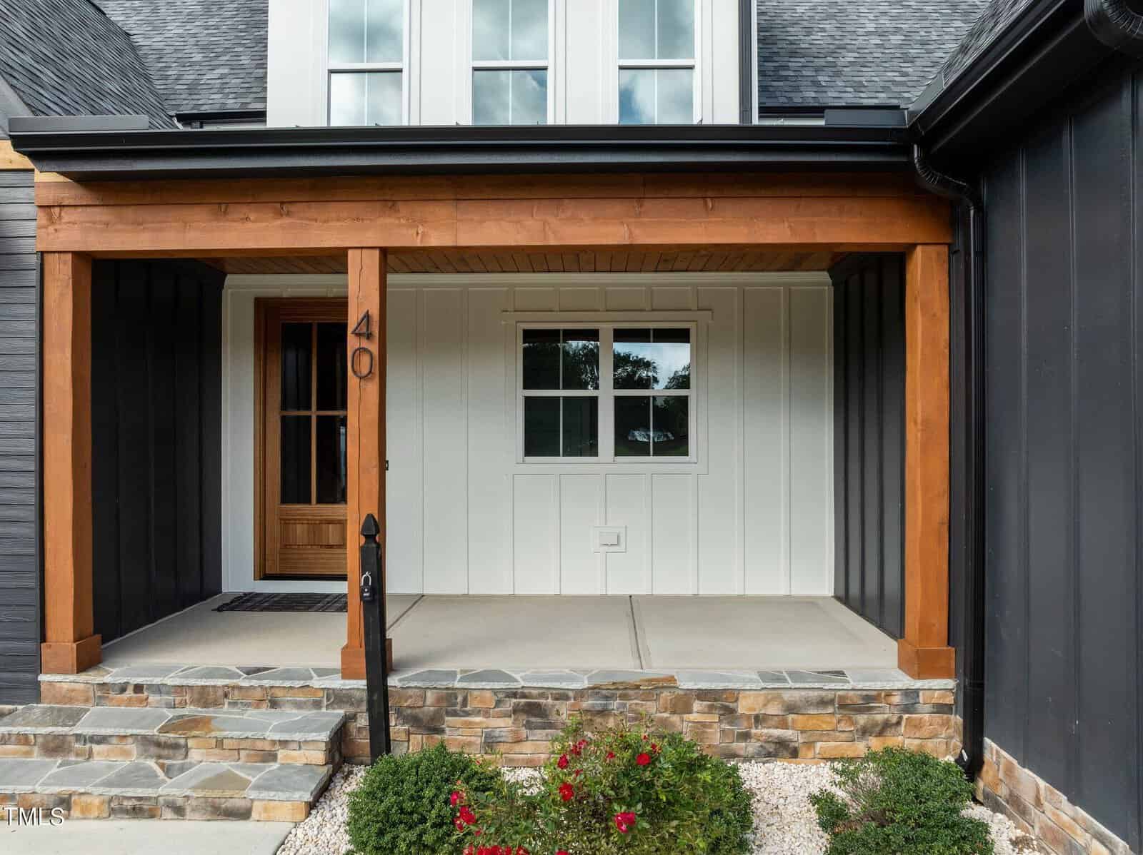 A modern house entryway with a wooden front door, white vertical paneling, black siding, a covered porch with wooden beams, stone steps, and small bushes with red flowers in front.