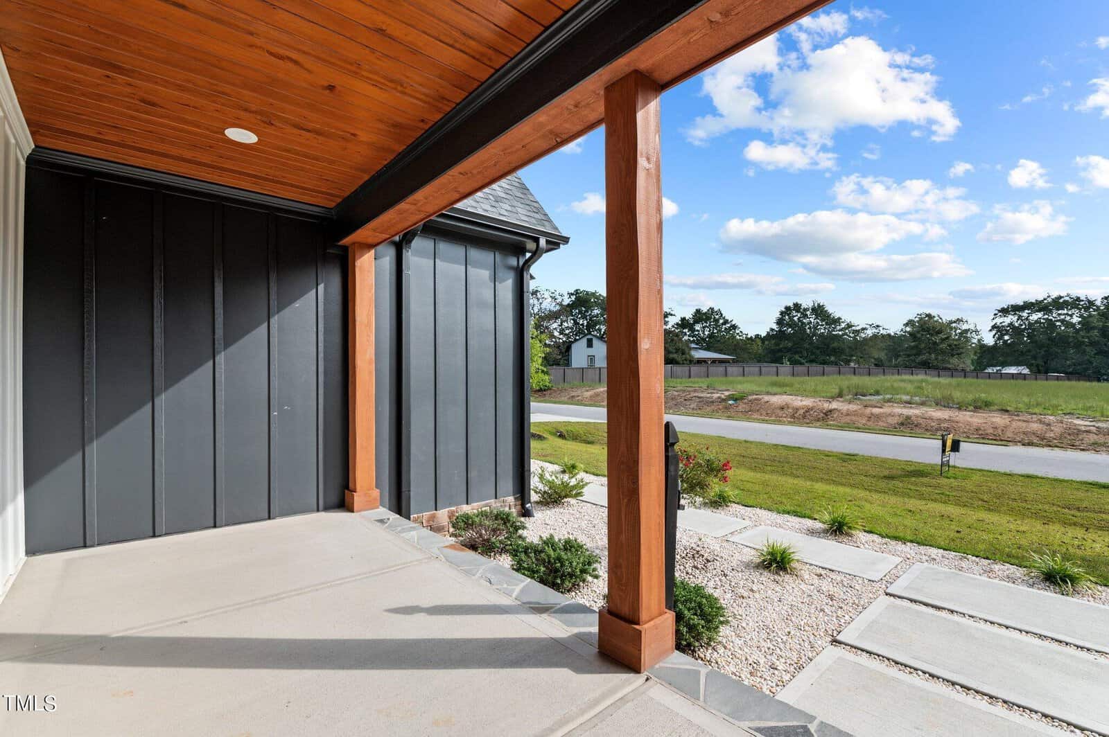 A modern house porch with wooden beams and a wood-paneled ceiling overlooks a landscaped front yard, a paved walkway, and a quiet suburban street with grassy fields and a few houses in the distance.
