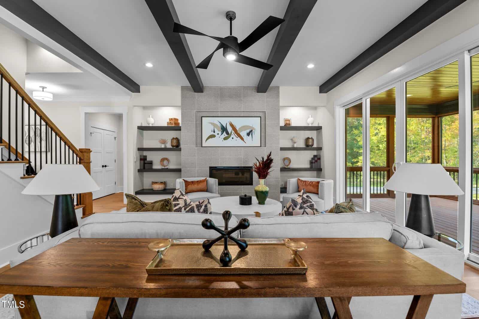 Modern living room with beamed ceiling, neutral decor, a gray stone fireplace, built-in shelves, large windows showing greenery, and a wooden console table behind a light-colored sofa.