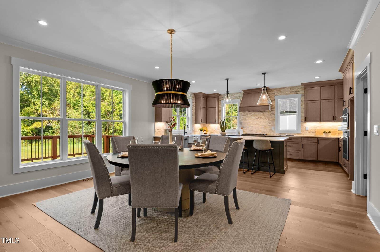 A modern kitchen and dining area with wooden floors, neutral-colored cabinets, a round dining table with six chairs, pendant lighting, large window, and an island with barstools. Lush greenery visible through the window.