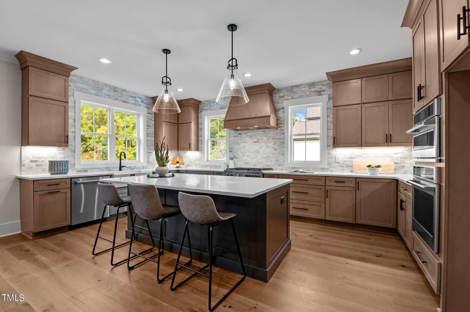 Modern kitchen with wood cabinets, a large black island with three barstools, pendant lights, stainless steel appliances, and two windows letting in natural light. Light wood floors and stone backsplash complete the space.