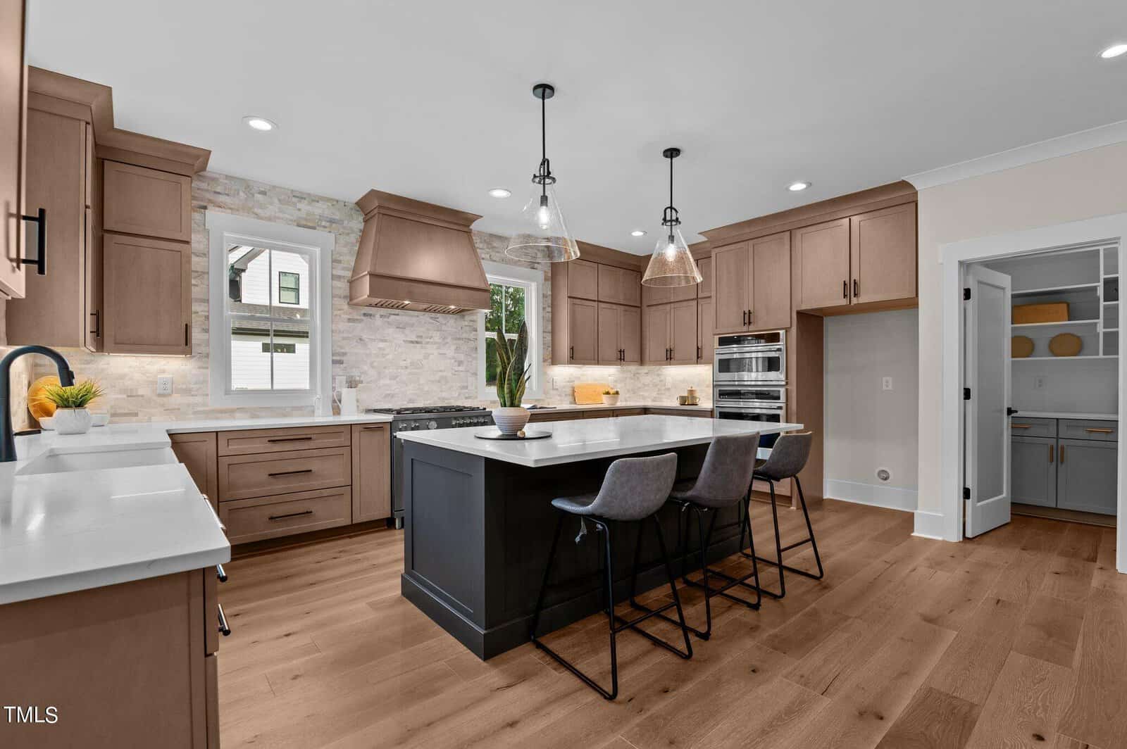 Modern kitchen with wood cabinets, white countertops, a large black island with three gray barstools, pendant lights, and hardwood floors. A window above the sink and a pantry with shelves are visible in the background.